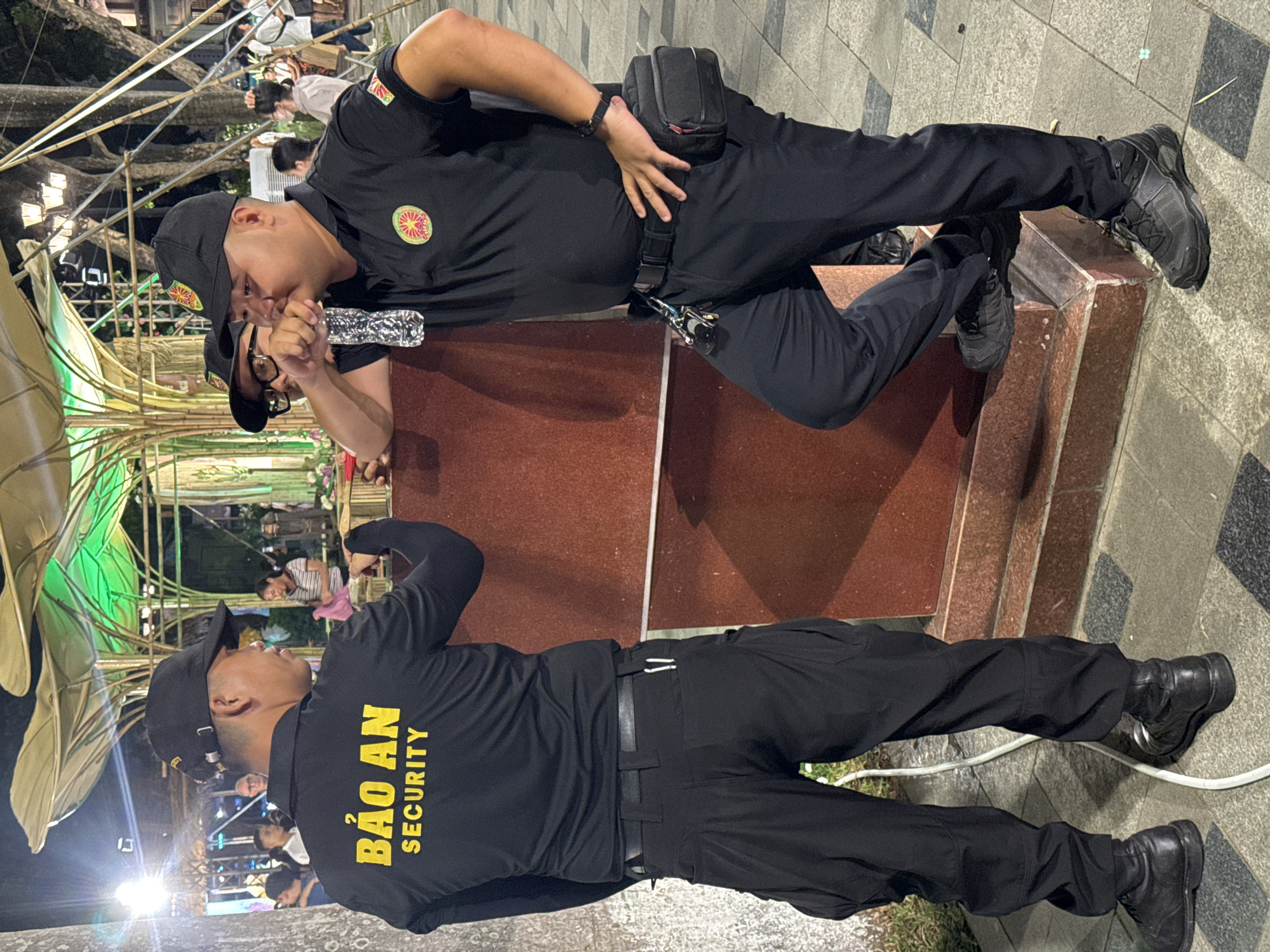 In this media file, two individuals are captured in a dimly lit outdoor setting, possibly at night, given the artificial lighting present. Both are dressed in black uniforms with the same circular emblem on their left breast and text on the back of their shirts. The individual in the foreground is standing and looking to the left. Their uniform displays the text "BẢO AN SECURITY" in yellow capital letters on the back. They are wearing black pants and black boots. The second individual is positioned behind and above the first, leaning on a reddish-brown structure. They are wearing a black cap and are holding a clear plastic bottle. Their uniform also bears the circular emblem.
The background suggests an urban environment, with elements of a building structure and possibly decorative lighting or installations, hinting at a public event or gathering. The presence of the security uniforms indicates that a security presence is likely in place. The overall mood appears casual, with the second individual seemingly taking a break and drinking from the bottle. The text on the uniform clearly identifies the organization as "BẢO AN SECURITY." The setting seems to be a public area in Hà Nội, Vietnam, as indicated by the location context. The lighting suggests it's either evening or nighttime.