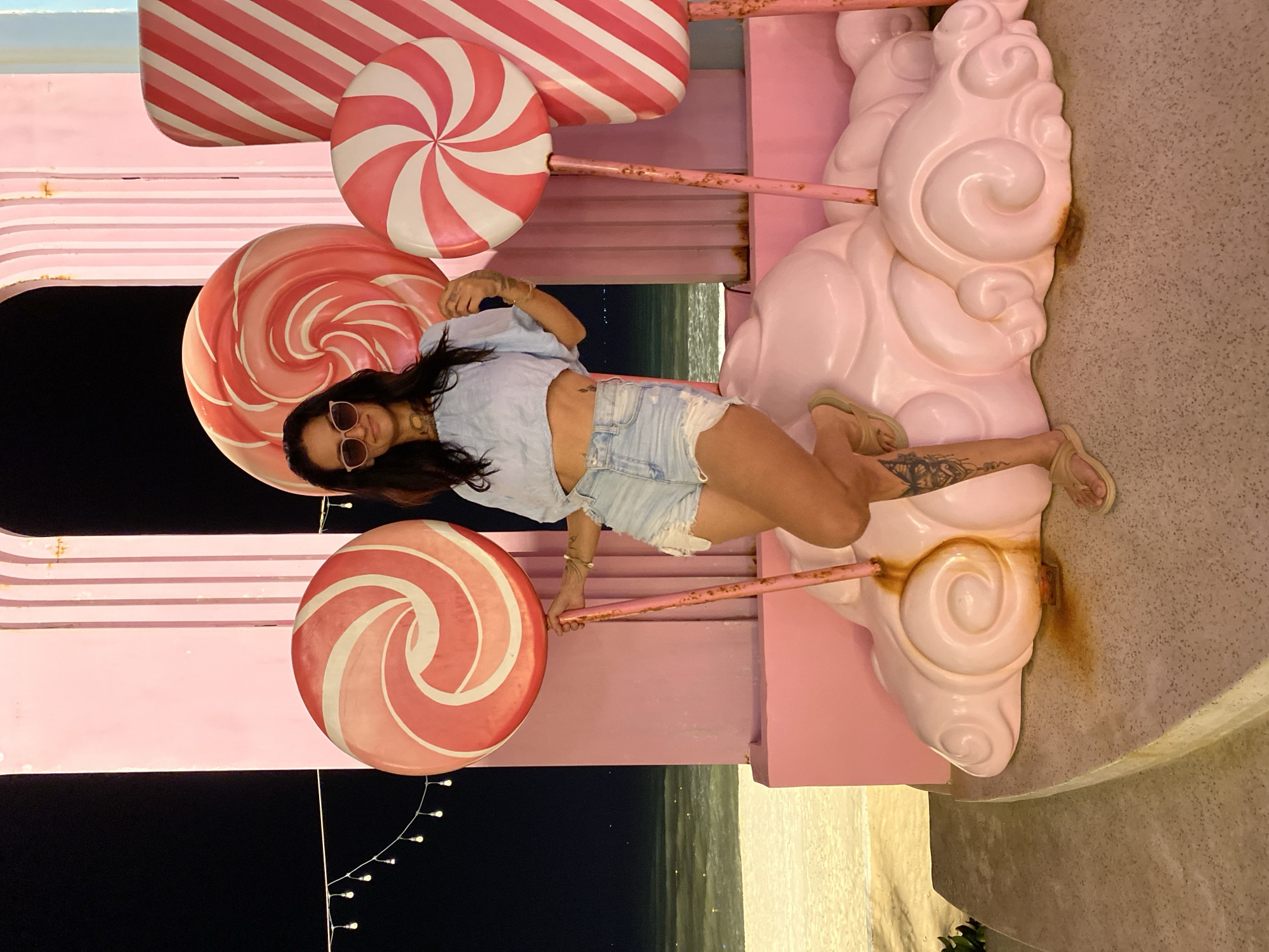 A woman is posing with giant lollipops in Tien Thanh Ward, Vietnam. She is wearing a light blue off-the-shoulder crop top and light wash denim shorts with frayed hems. She has sunglasses on and her dark hair is down. Her right leg is bent, with her foot resting on a pink cloud-like structure. She has tattoos on her left leg. Behind her are several oversized, brightly colored lollipops and pink architectural elements. The scene appears to be set up for photography or decoration, possibly at a resort or attraction. The background shows a glimpse of the ocean and a beach at night, with string lights visible in the distance. The ground in the foreground is concrete with a textured finish.