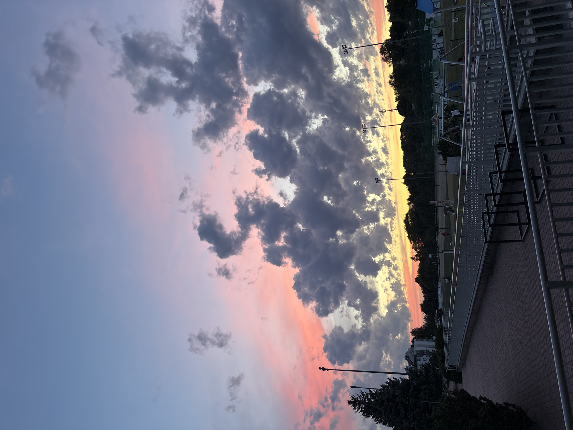 This is a landscape photograph taken at sunset in Lublin, Poland. The scene captures a sky filled with dramatic clouds tinged with pink and orange hues from the setting sun, contrasted with darker, gray clouds. In the foreground, a paved walkway made of reddish-brown bricks is visible, bordered by a silver metal railing. Beyond the walkway, a sports field or recreational area is present, with sports lighting poles and trees lining the horizon. A building, possibly residential, is seen to the left of the frame, partially obscured by a large evergreen tree. The overall atmosphere is serene, with the vibrant colors of the sky dominating the composition.