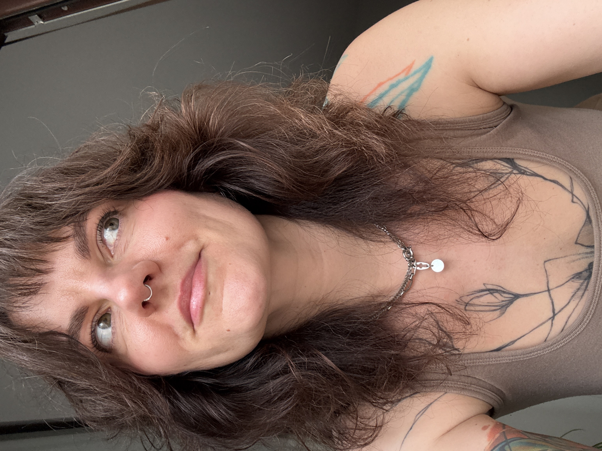 A woman with brown, wavy hair and a septum piercing in her nose is shown from a low angle, looking slightly upward and to the left. She is wearing a brown tank top and a silver chain necklace with a small circular pendant. Tattoos are visible on her upper chest and shoulder. The background is a plain, gray wall. There is no information in the image indicating a location of Boreti, Montenegro.