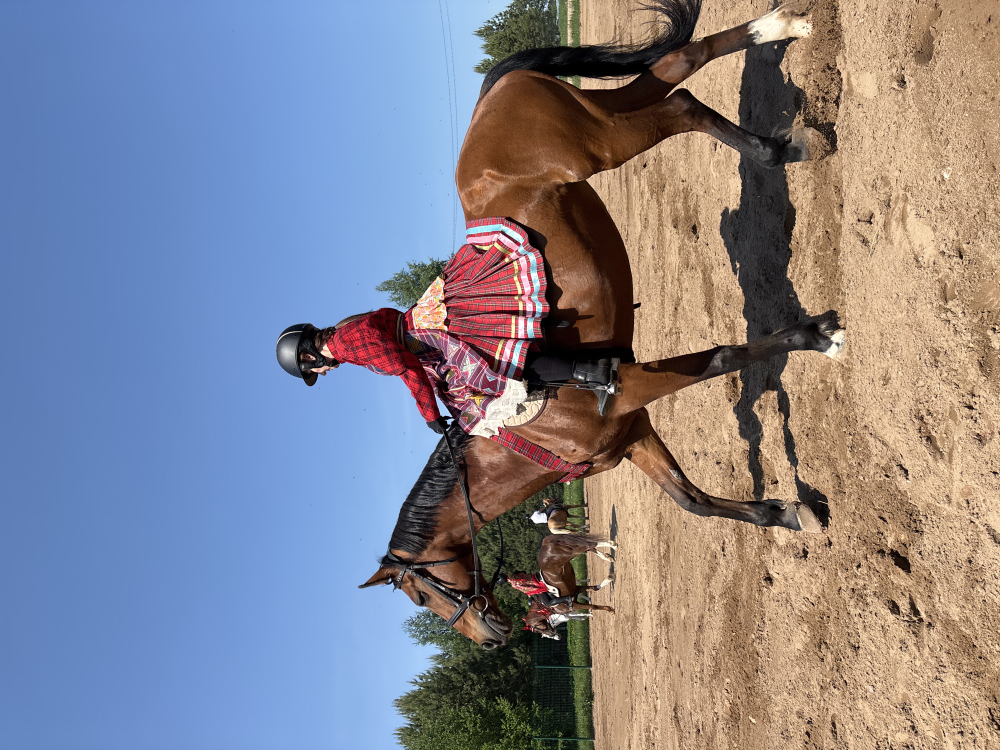 The image depicts a person riding a brown horse in an outdoor sandy arena. The rider is wearing a black helmet, a red plaid top, and a large red plaid skirt or drape over the horse's back. The horse is equipped with a bridle and saddle. In the background, additional horses and riders can be seen. The environment includes trees, a blue sky, and what appears to be a fenced area. The location is Русский Вожой, Russia.