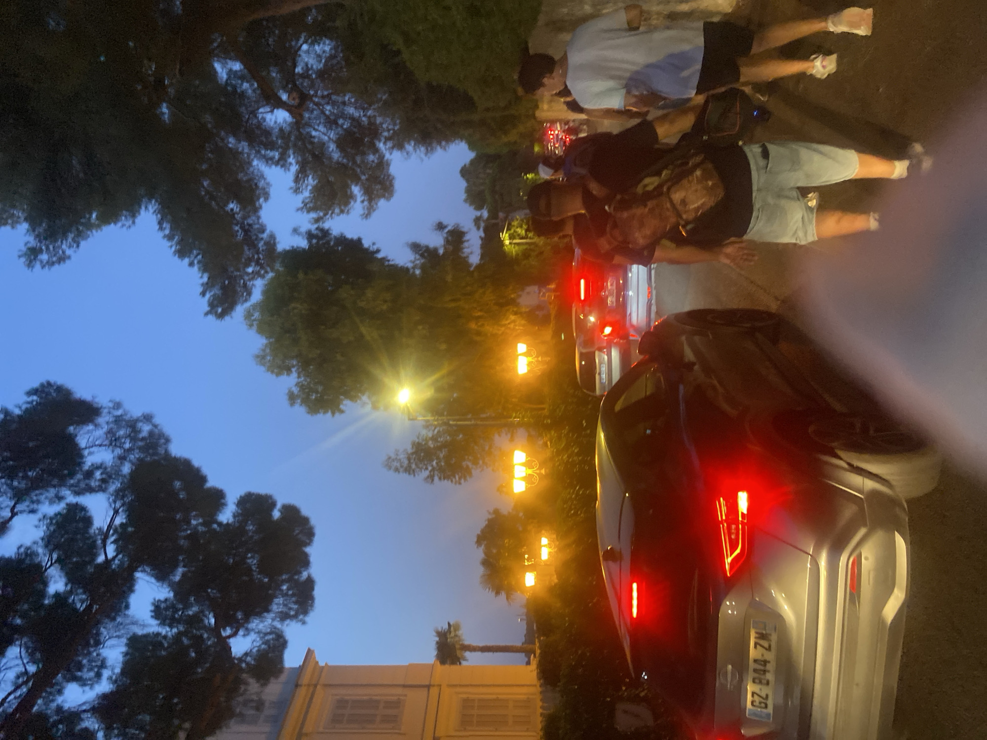 In the city of Antibes, France, at dusk, a street scene unfolds with traffic congestion and pedestrians. A gray Nissan SUV with illuminated red taillights is prominent in the foreground, its license plate reading "GZ-B44-ZM." Several individuals are walking along the street, with one person in the middle ground wearing a camouflage backpack and light blue shorts. To the right, another person in a light blue t-shirt and dark shorts walks with white athletic shoes. In the background, streetlights cast a warm glow, illuminating palm trees and the facade of a building with distinctive window shutters. The sky is a deep twilight blue.