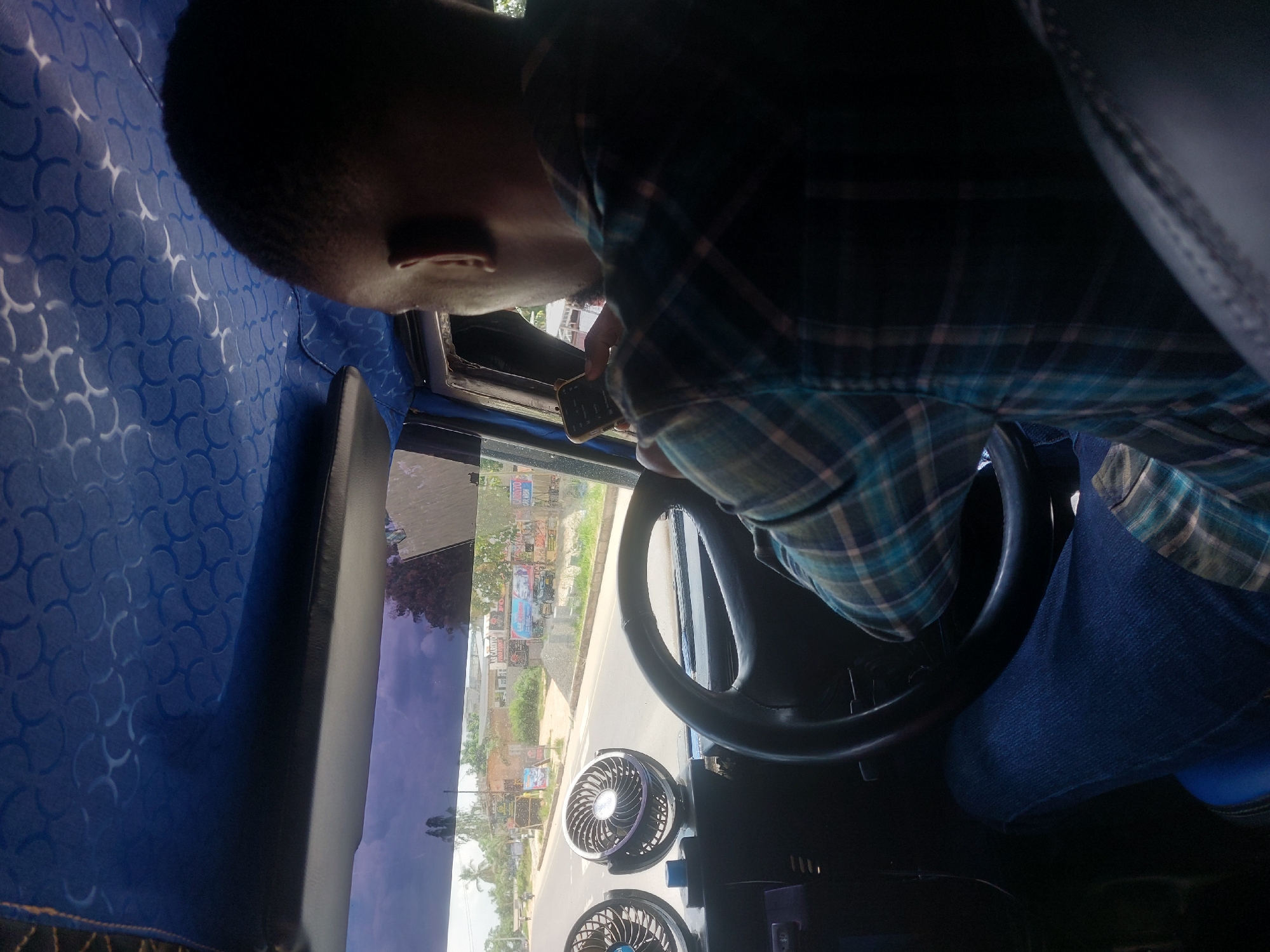 The image, taken from within the vehicle, shows a person driving a car on a road in what appears to be Dar es Salaam, Tanzania. The driver is glancing at a phone in their left hand. The vehicle interior is predominantly blue with a distinct patterned ceiling. Two small, battery-operated fans are visible on the dashboard. The view through the windshield shows a street lined with buildings and billboards.