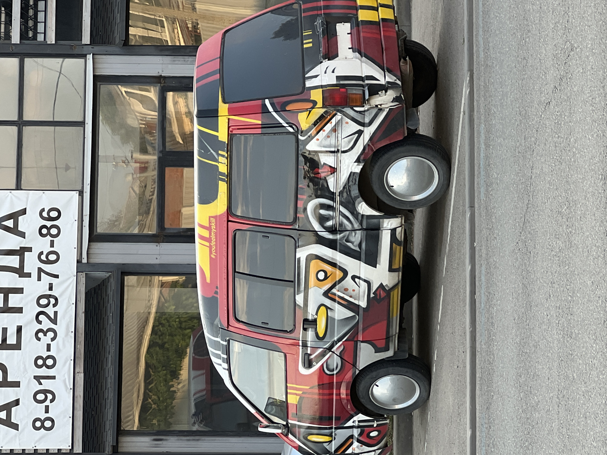 This media file shows a brightly painted van parked next to a building with large windows. The van is decorated with a colorful, abstract mural featuring red, yellow, black, and white patterns. The mural has a graffiti-like style with sharp lines and geometric shapes. Visible on the side of the van is a hashtag: "#youfeelmyskill". The building behind the van appears to be a commercial establishment, possibly an office or retail space, with reflective windows. A white banner with black text is partially visible on the left side of the image. The text on the banner is in Russian and appears to be an advertisement for rental services, with the visible portion reading "АРЕНДА 8-918-329-76-86", which translates to "RENTAL 8-918-329-76-86". The scene is outdoors, with asphalt visible in the foreground and on the right side of the frame, suggesting a street or parking area. The lighting suggests it is daytime, and the overall atmosphere is neutral. The image is oriented vertically, with the van and building appearing on their sides.