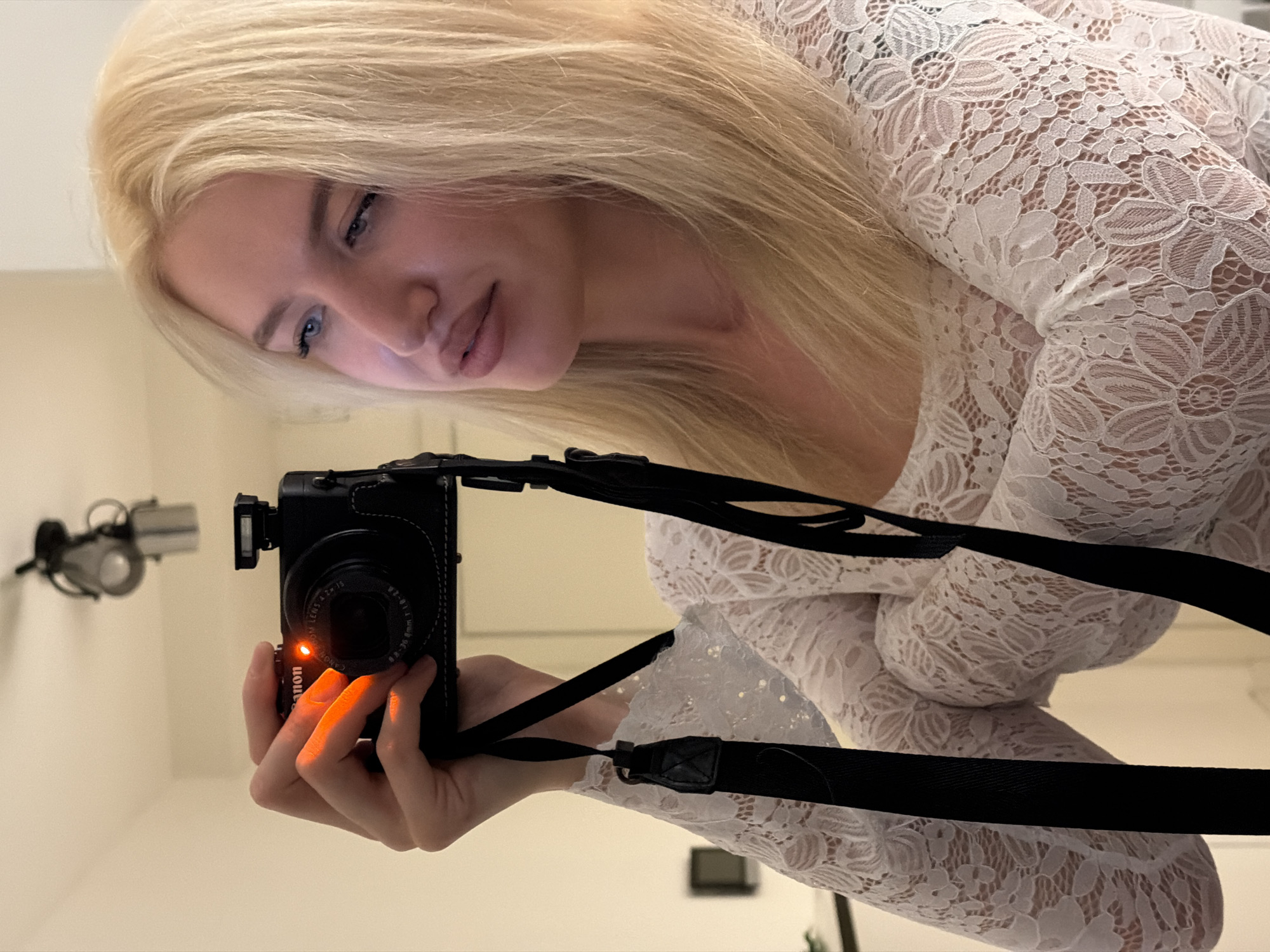 An indoor scene depicts a mirror selfie of a female individual in Belgrade, Serbia. The subject, a woman with long blonde hair, fair skin, and blue eyes, is captured from the chest up, looking slightly to her right with a neutral expression. Her left hand holds a black Canon compact digital camera, identifiable by "Canon" branding and lens specifications (ZOOM LENS 8.8-36mm 1:1.8-2.8), pointed towards a reflective surface. A prominent red indicator light is illuminated on the front of the camera. A black camera strap is visible, hanging from the device. The woman is wearing a white, long-sleeved lace top featuring an intricate floral pattern. The background consists of a plain, light-colored wall. A ceiling-mounted spotlight fixture with two directional lamps is visible in the reflection above and behind the camera. The scene is illuminated by warm, artificial light.