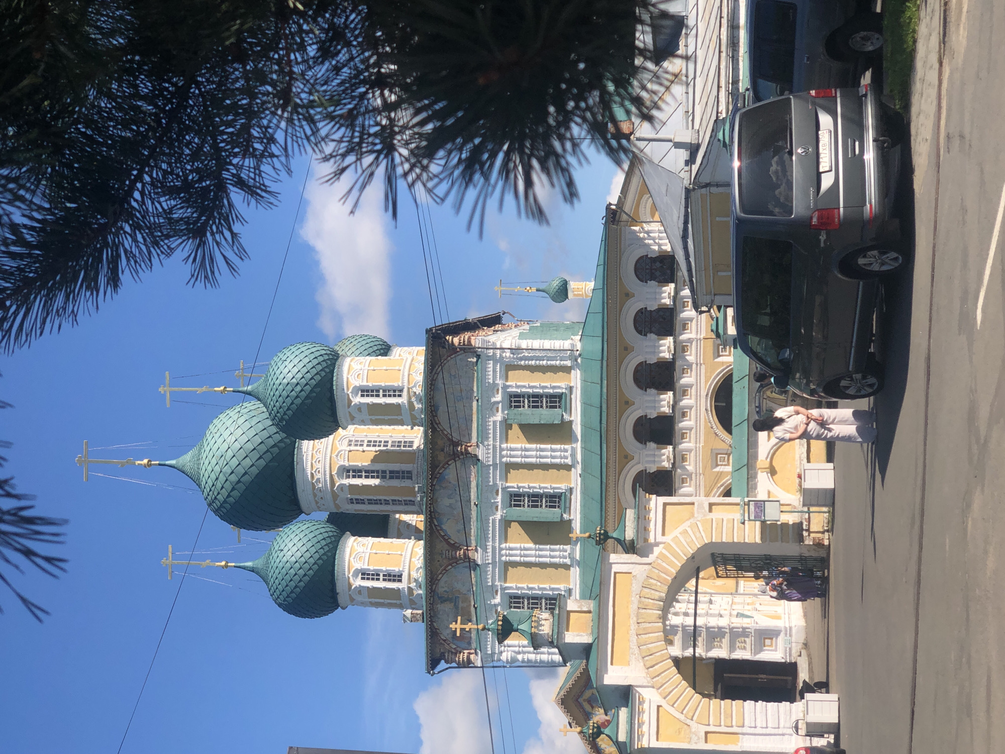 The image shows a sunny day in Tutaev, Russia, with the Voskresensky Cathedral prominently featured. The cathedral, a yellow and white building with teal domes topped with golden crosses, is partially obscured by a dark green pine tree in the foreground. Several people are visible near the cathedral's arched entrance: two individuals in purple robes stand by a gate, while another person in white stands facing away from the camera in the foreground. Two vehicles, a dark gray minivan and part of a blue SUV, are parked on the asphalt road. The sky is a clear blue with a few scattered white clouds.