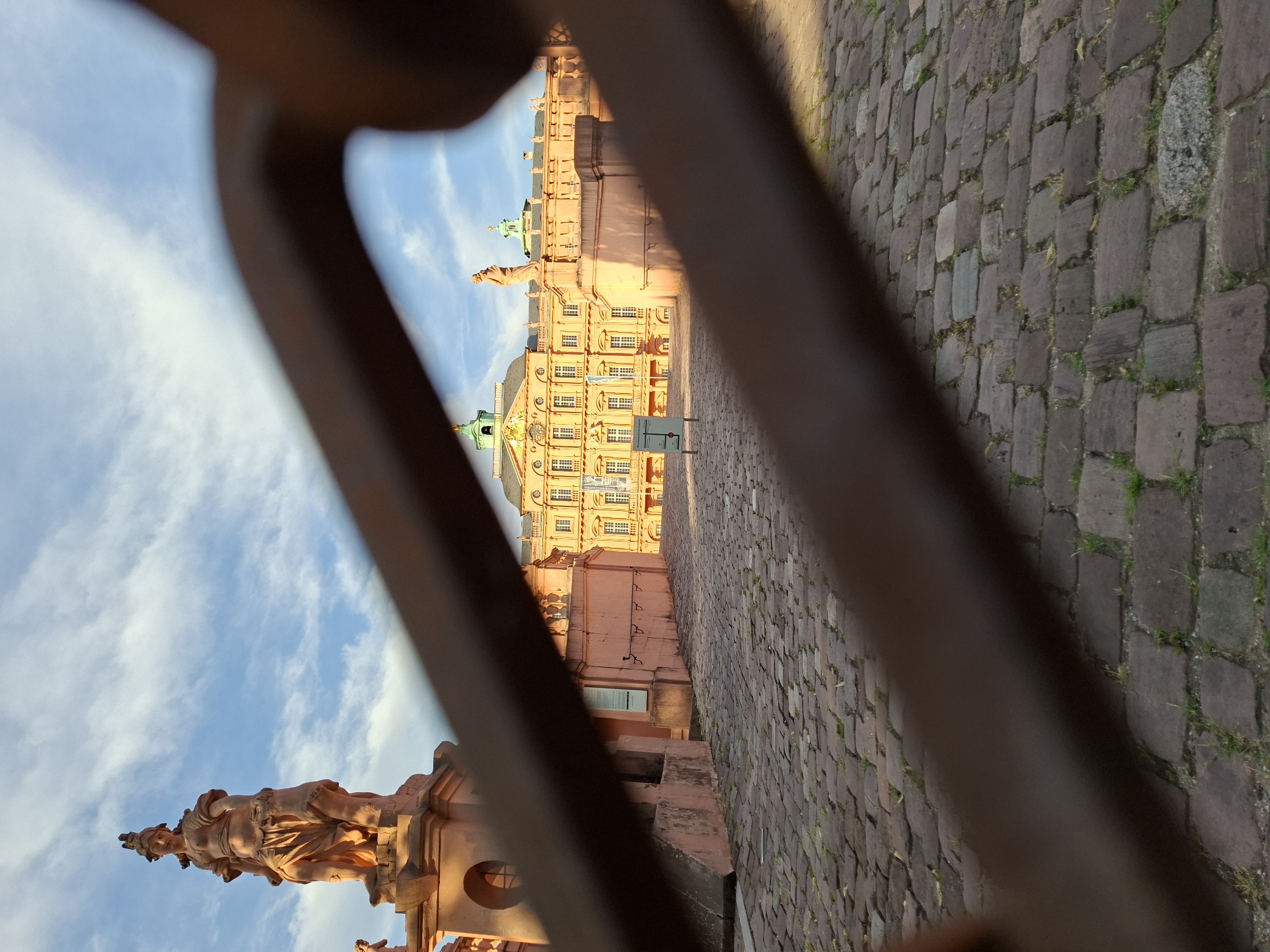 The image, taken from a low angle, shows a cobblestone path leading towards a pink Baroque palace. In the foreground, a blurred, dark brown metal railing is prominently featured, partially obscuring the view of the palace and its surroundings. On the left side of the path, a stone statue of a woman in classical attire stands atop a pedestal. The palace, with its numerous windows and ornate architectural details, is illuminated by the golden light of what appears to be late afternoon or early morning sun. The sky above is blue with scattered white clouds. The scene is located at the Favorite Palace in Rastatt, Germany, a historic building known for its Baroque architecture and beautiful gardens.