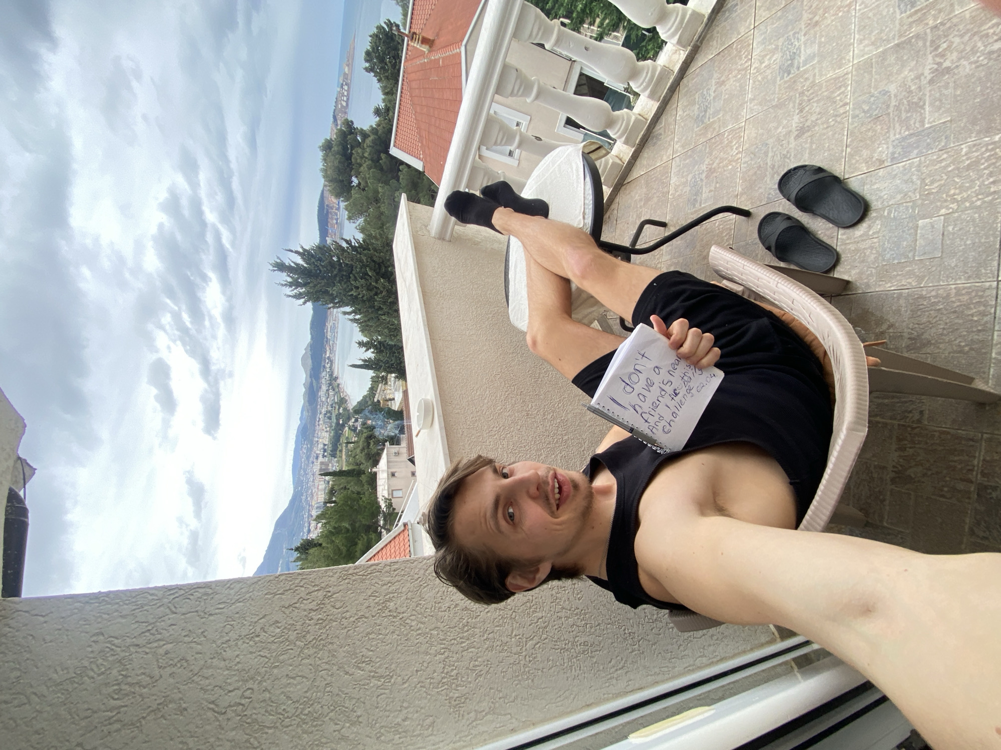 A male person is taking a selfie on a balcony. He is wearing a black tank top and black socks, and is sitting with his legs crossed on a small white table. He is holding a notebook with writing on it. A pair of black flip-flops sit on the tiled balcony floor. The balcony overlooks the coastal city of Šušanj, Montenegro, with mountains in the background. The sky is overcast. The balcony has a white balustrade and is attached to a building with a red-tiled roof.
