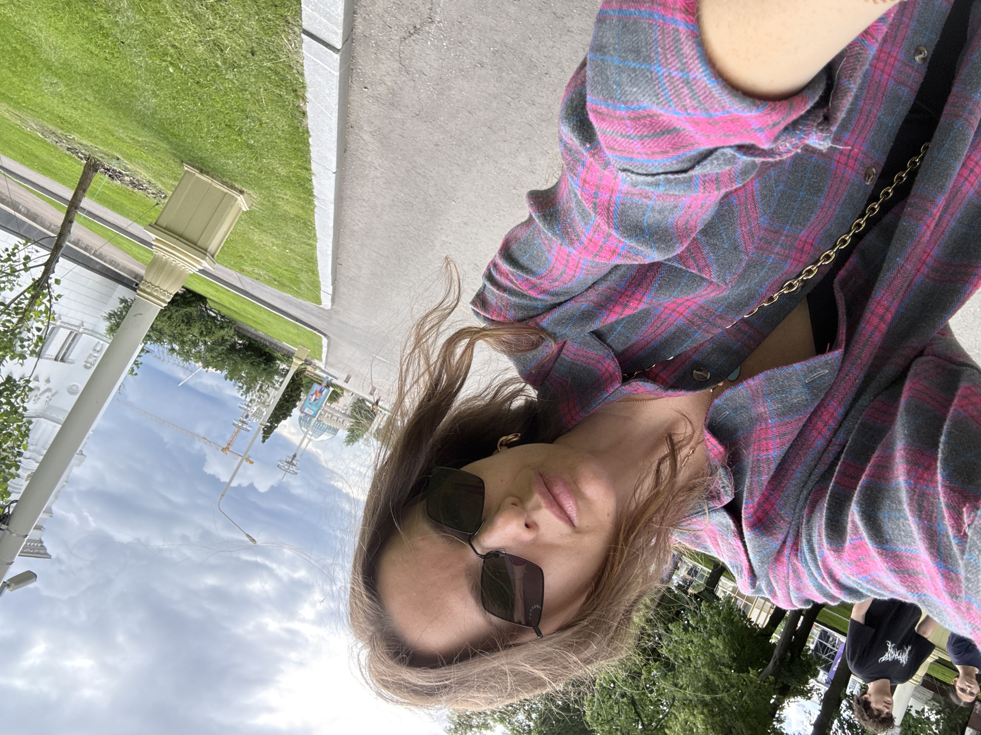 A woman is taking a selfie outdoors on a cloudy day. She is wearing a pink and gray plaid shirt, sunglasses, and a gold chain. In the background, there are trees, a street, grass, a building with classical architecture, a lamppost, and a construction crane. Two other people are visible in the background, one wearing a black t-shirt with white lettering. The location is VDNKh (Exhibition of Achievements of National Economy) in Moscow, Russia, specifically near the "Cosmos" Pavilion.
