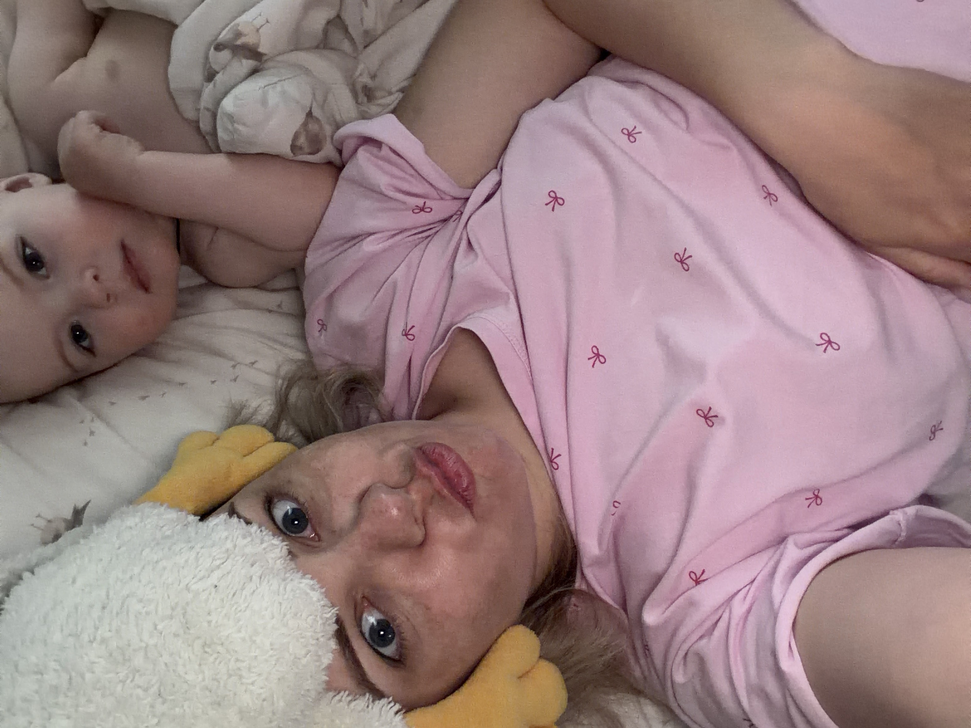 A woman and a baby lie on a bed. The woman is wearing pink pajamas with bow patterns and is partially covered by a white and yellow stuffed animal. The baby is near her head on the left side of the image. The location is unknown, but it is not in Votkinsk, Russia.