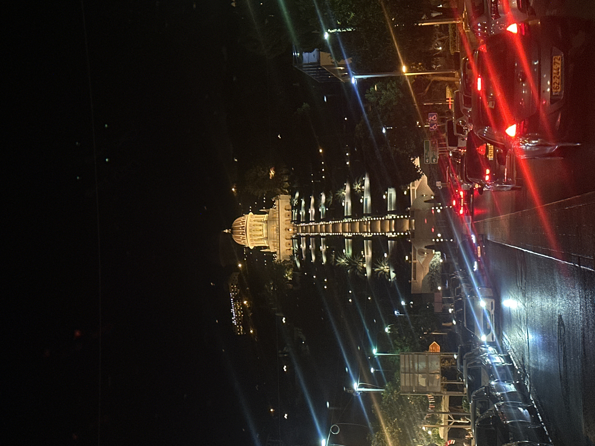 This is a night-time photograph taken from a vehicle, likely a car or bus, showing a view of a building and a street with cars. The image is rotated, with the street and building appearing vertical.
The main subject is a large, ornate building with a domed roof, illuminated by numerous lights. The building appears to be a place of worship or a significant landmark. It is surrounded by trees and other structures, also lit up. In the foreground, a line of cars is visible on a wet street, their headlights and taillights reflecting on the road. The reflections suggest it might have recently rained or the road is being washed.
The activity taking place seems to be a nighttime journey or observation of the city. The presence of cars on the road indicates normal traffic flow, even at night. The atmosphere is somewhat serene and picturesque due to the lighting of the buildings and reflections on the wet street.
Notable details include the time of day, which is clearly night, indicated by the dark sky and artificial lighting. The weather appears to be damp or rainy, based on the reflections on the road. Location cues are provided by the license plates of the cars, which are in Hebrew script and appear to be from Israel. One license plate clearly visible reads "62-244-72". Another reads "573-13". There are also road signs with Hebrew text.
The overall impression is that of a beautiful nighttime scene in Haifa, Israel, with a prominent illuminated building as the focal point, set against the backdrop of a city street at night.