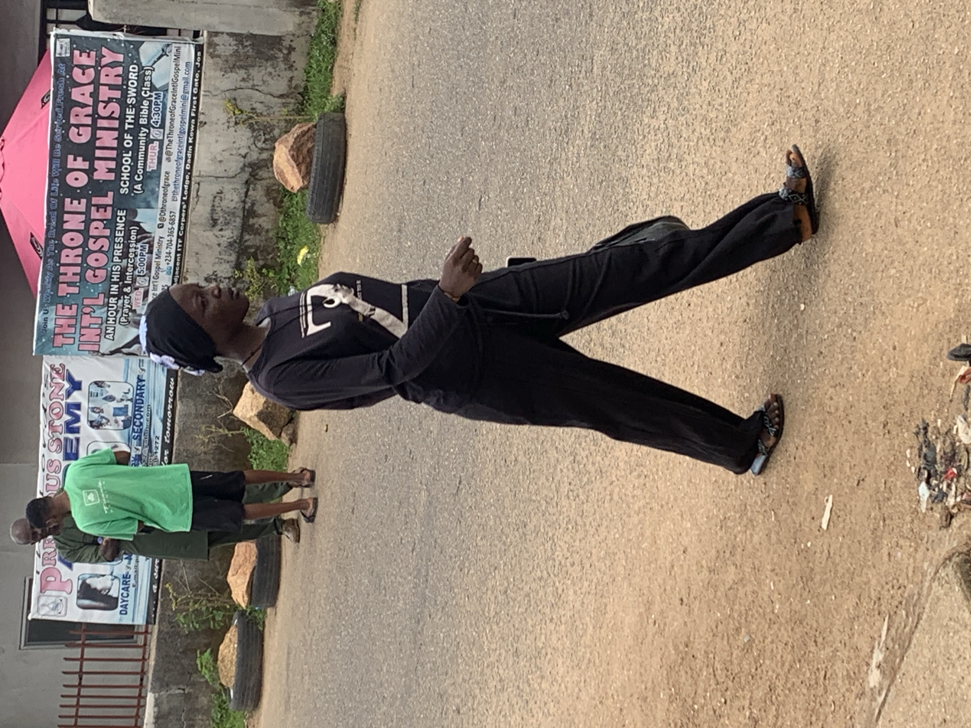 In Jos, Nigeria, on a paved road, a Black woman wearing a black headscarf, black long-sleeved shirt with a graphic print, and black pants walks from left to right. She is wearing sandals. Behind her, two Black men are talking, one in a green t-shirt and shorts, and the other in a dark green robe. A banner behind them reads "PRECIUS STONE ACADEMY" and "THE THRONE OF GRACE INT'L GOSPEL MINISTRY". The banner also lists service times and contact information, including a phone number and email address. The environment includes a concrete barrier, some rocks, and discarded tires. Garbage is visible on the side of the road.
