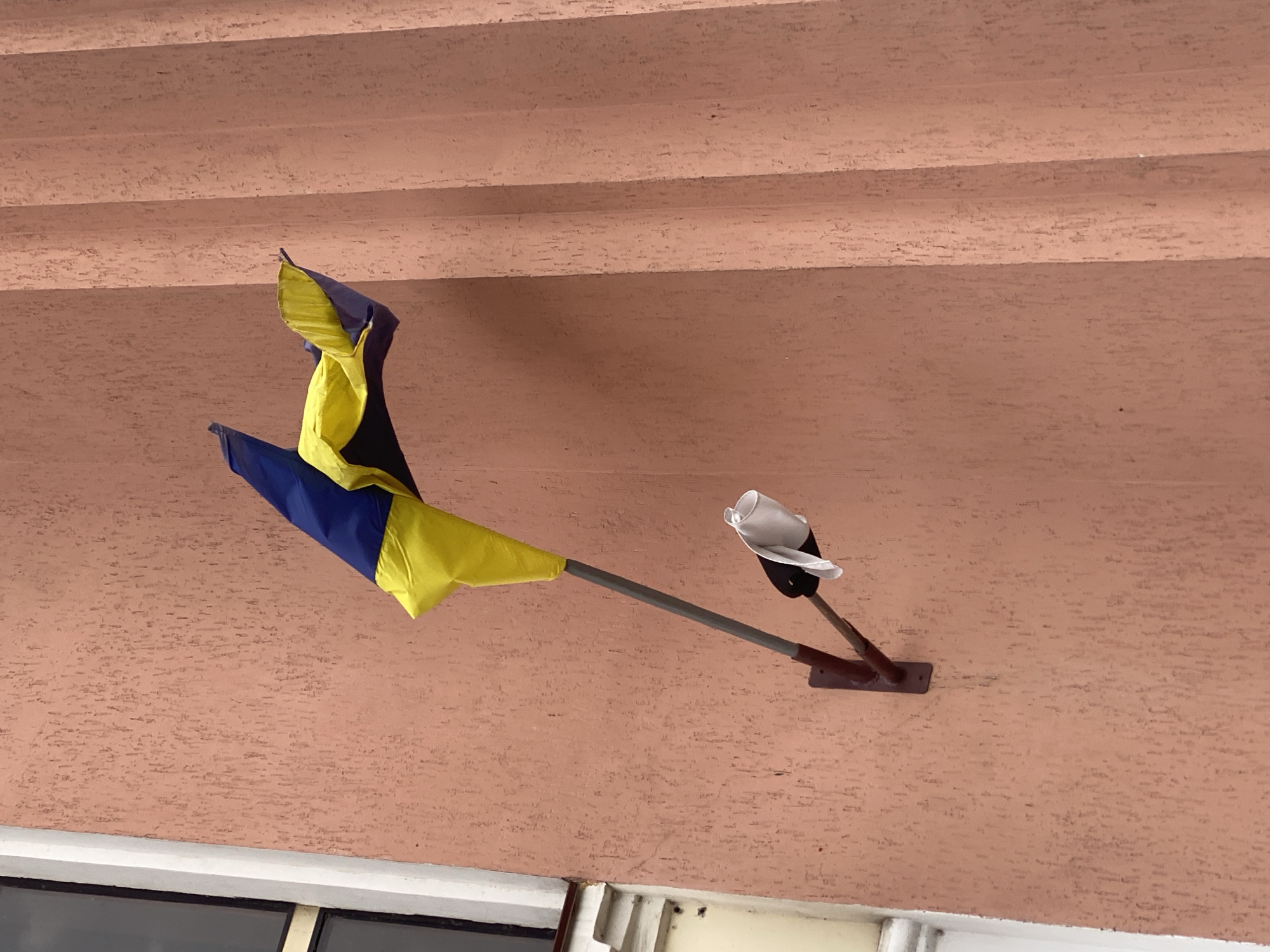 A Ukrainian flag, consisting of blue and yellow horizontal stripes, is affixed to a pole extending from a textured, peach-colored wall. The flag is partially furled and appears somewhat tattered, with its lower section draped over the pole. Attached to the pole below the flag is a smaller, white cloth object, possibly a handkerchief or a piece of paper, also appearing somewhat crumpled. The wall itself has a rough, stucco-like finish and is vertically ribbed by what appear to be architectural columns or decorative elements. To the left of the flag assembly, a portion of a window with white trim is visible, suggesting the flag is mounted on the exterior of a building. The lighting suggests it is daytime, with diffused light casting soft shadows. This scene is located in the city of Stadnia, Ukraine.