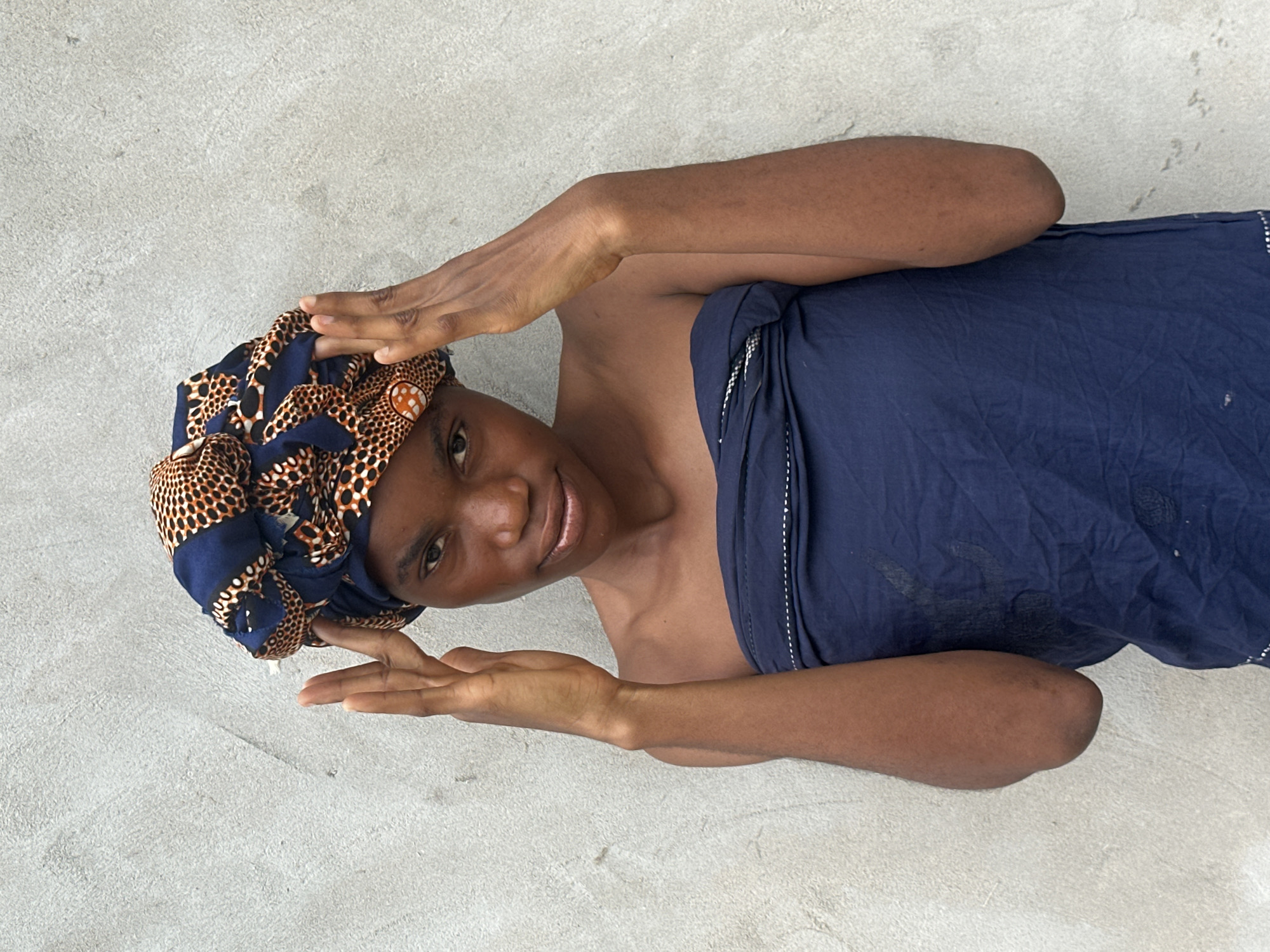 A young person, appearing to be a girl, lies on a textured, light-colored surface that resembles concrete. She is wearing a headwrap with a dark blue and orange patterned fabric, and a dark blue garment that covers her torso and extends downwards. Her hands are positioned on either side of her head, palms facing outwards, and she is looking towards the camera with a slight smile. The lighting suggests it is daytime, possibly outdoors, with soft light. The background is a plain, textured surface, with no other objects or distinct features visible. The overall impression is one of a posed portrait. Given the location context of Badagry, Nigeria, the scene could be interpreted as a casual moment captured at home or in an outdoor public space.