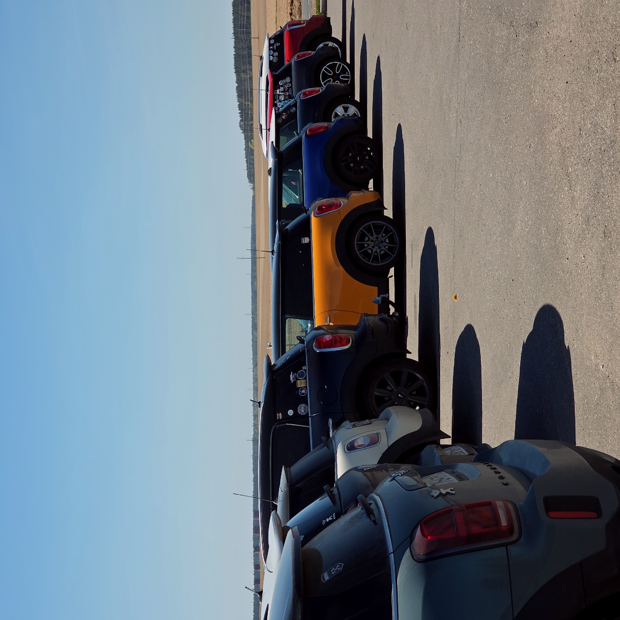A row of Mini Cooper automobiles, primarily viewed from the rear, are parked on an asphalt surface under a clear, light blue sky. The cars are positioned lengthwise in the frame, and based on the direction of the shadows, it is likely daytime. The cars are a variety of colors, including red, black, blue, orange, white, and green. The background shows a distant treeline and a flat plain. The location is Свіслацкі сельскі Савет, Belarus.