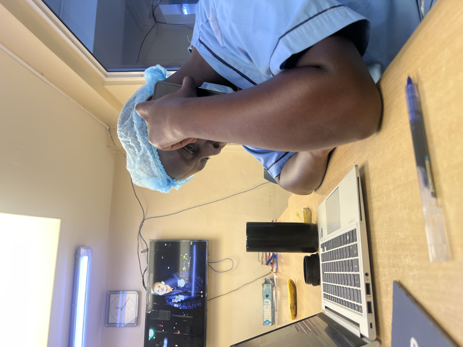 An individual is seated at a light-colored wooden desk in an indoor room, identified as a location in Nairobi, Kenya. The person, appearing to be an adult with dark skin, is wearing light blue medical scrubs and a matching blue disposable surgical cap. A face mask hangs below their chin. They are holding a black smartphone to their right ear with their right hand, engaged in a conversation. Their left arm is bent, resting on the desk. On the desk in the foreground, a silver laptop is open, a black cylindrical container (possibly a thermos) with its lid removed sits next to it, and a yellow banana is visible behind the laptop. A blue pen and a partially visible blue folder are also on the desk. In the background, a flat-screen television mounted on a light beige wall displays a static image of a young person. Above the television, a square analog wall clock is visible, alongside a long fluorescent light fixture on the ceiling. Black electrical cables are present along the wall. To the far right, a window or glass panel shows a muted blue light, indicating an external view.