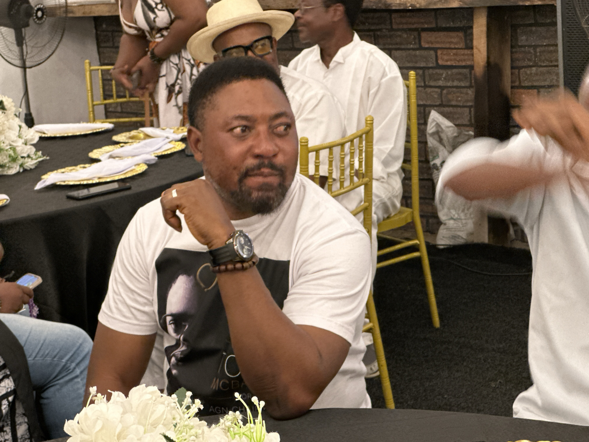 In Barden Yana, Nigeria, a man with dark skin, a goatee, and a black t-shirt with a portrait on it is seated at a black table, his right hand resting on his chin. He wears a watch on his left wrist and a ring on his right hand. To his left, a woman in a white dress with a floral pattern stands. Behind him, a man in a white shirt and hat is visible, and further back, another man in a white shirt is seated. The table is set with gold plates and white napkins. A large fan is positioned to the left of the frame, and a brick wall is visible in the background. The lighting suggests an indoor evening event.