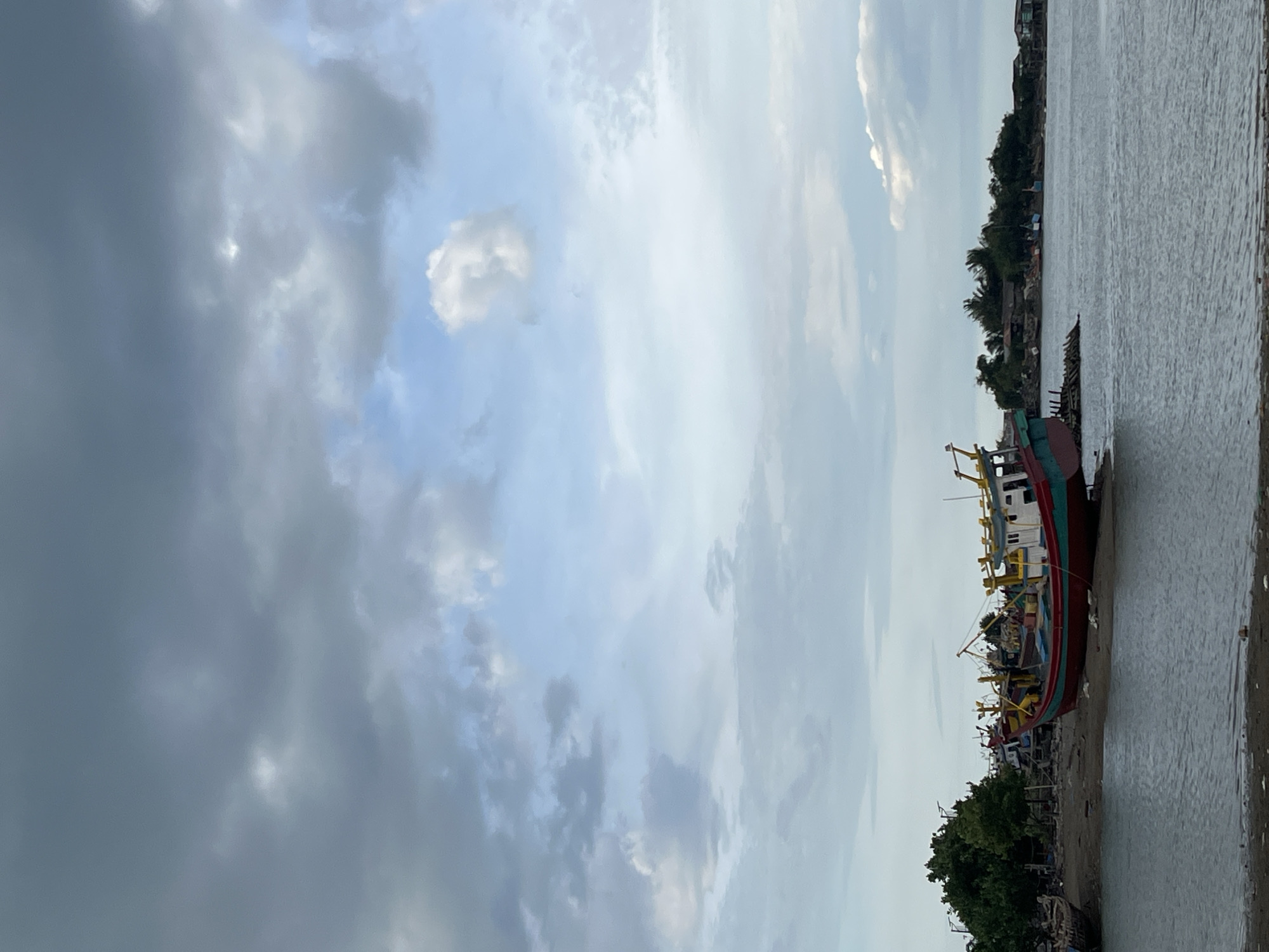 The image depicts a coastal scene in Lhokseumawe, Indonesia. The sky occupies a significant portion of the image, exhibiting a mix of dark, overcast clouds on the left and patches of lighter, blue sky with scattered clouds on the right. Below the sky, a body of water, likely a river or bay, stretches horizontally across the frame. Along the shoreline to the right, there's a sandy or muddy bank where a large, multi-colored fishing boat is beached. The boat is predominantly red and green, with visible yellow and white details. Trees and vegetation grow densely along the shore, and a few buildings are visible in the background, suggesting a populated area. The overall scene appears calm, with no people immediately visible in the frame.