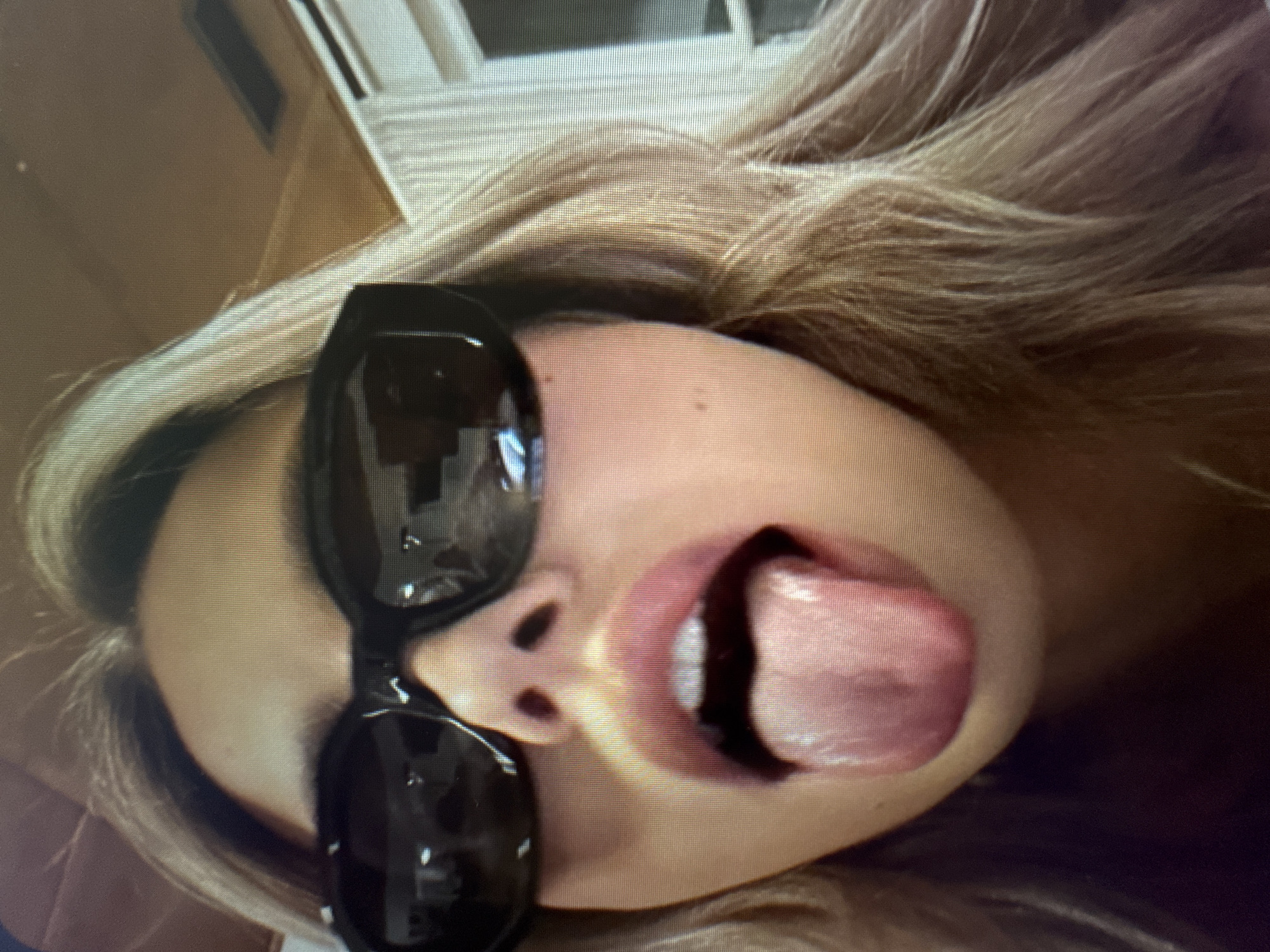 The image displays a close-up, rotated view of an individual's face, captured with visible scan lines and pixelation, suggesting it is a photograph of a digital screen. The person's head is tilted approximately 90 degrees clockwise. They are wearing large, dark-framed sunglasses, which reflect a blurry indoor environment, possibly a ceiling or window. Long, light brown or blonde hair, styled with some waves, frames their face. Their mouth is wide open, with a pinkish tongue extended outward in a playful or cheeky gesture, revealing a hint of white teeth on the visible side. The bridge of their nose is visible between the sunglasses and mouth. The skin tone appears light, with a few faint blemishes visible near the hairline in the cropped hints. The background is indistinct but appears to be an indoor setting. No specific location, time of day, or visible text can be determined.