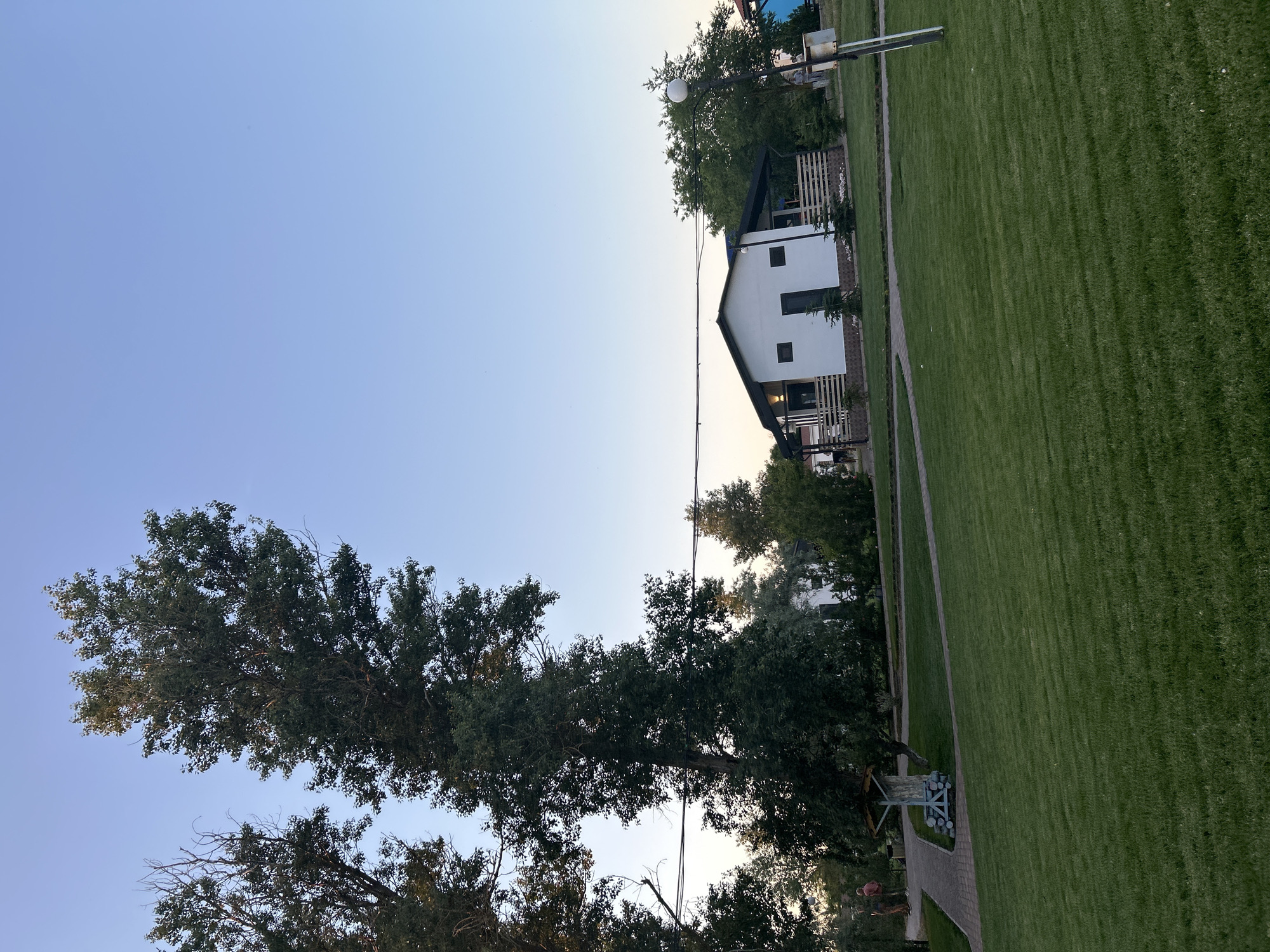 The image depicts a clear, pale blue sky over a well-maintained grassy area in Останинское сельское поселение, Ukraine. In the mid-ground, a modern white house with a dark roof is visible, featuring a wooden fence and several small trees. To the left of the house, two tall trees with dark green foliage dominate the left side of the frame, with one tree extending to the top edge of the image. A paved pathway winds through the lawn, leading towards a small, rustic wooden structure. In the far left of the image, two individuals are walking on the pathway. A lamppost with a spherical light fixture stands on the right side of the image. The overall scene suggests a serene, residential or recreational outdoor setting.