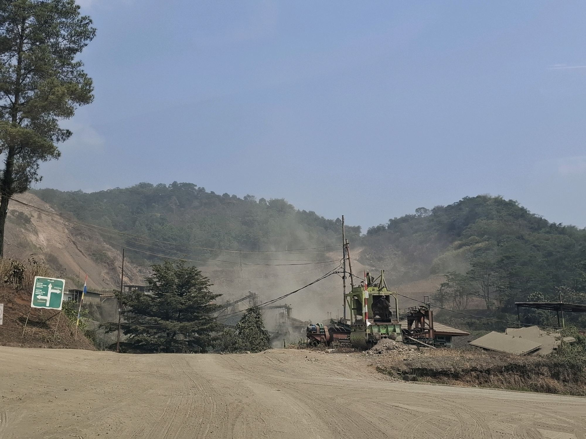This is a medium-shot, eye-level, outdoor, daytime photograph of a mining or construction site. In the foreground, a dirt road is visible with tire tracks. To the left, a signpost with green directional arrows and text in Indonesian points to the left and right. Beyond the road, a mining or processing facility is active, with machinery and structures, including a large green machine with red and white striped accents, conveyor belts, and buildings. Dust is rising from the site, obscuring some of the machinery and the background. In the middle ground, several trees, including a tall pine tree on the left, are present. The background consists of lush, green, tree-covered hills and mountains under a clear, pale blue sky. The environment appears to be a hilly, possibly tropical or subtropical region. The scene conveys an industrial operation in a natural landscape. The specific location is the city of Cijangkar, Indonesia, as indicated by the signpost.
