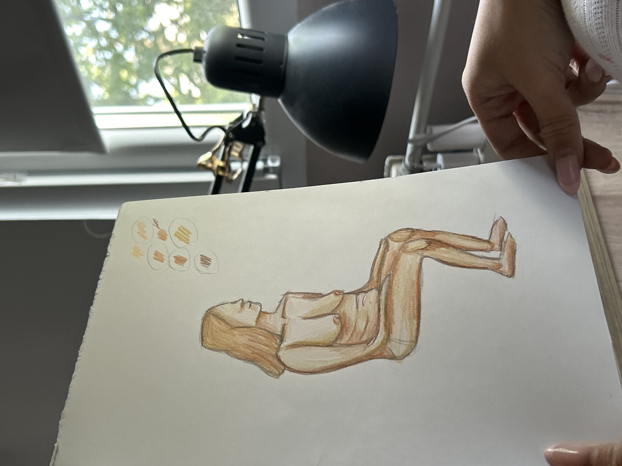 A drawing of a nude figure is held by a person with manicured nails. The drawing is lit by a black adjustable desk lamp. The background includes a window with blinds and greenery visible outside. The location is not discernible from the given image.