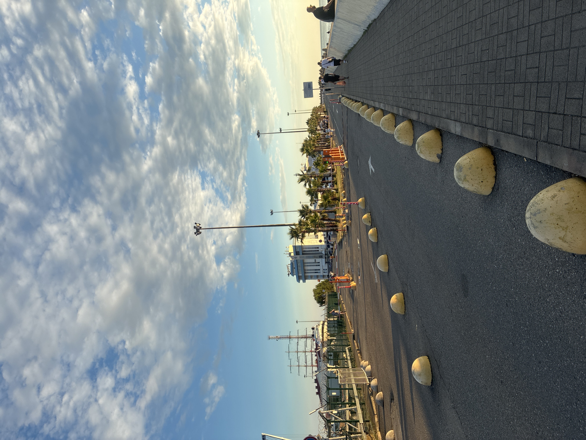This is a vertically oriented, eye-level, medium shot of a paved walkway and road, likely at a seaside location given the palm trees and ship in the distance. The sun is low in the sky, casting long shadows and illuminating the clouds with a golden light, suggesting it is either sunrise or sunset. The sky is a mix of blue and white clouds, with some appearing wispy and others more solid.
On the left side of the image, a wide expanse of clouds dominates the sky, with patches of blue peeking through. A tall lamppost with multiple lights is visible. In the distance, a large, white building with several windows is situated amongst palm trees. Further in the background, a docked sailboat with multiple masts is prominent.
The walkway, made of dark grey paving stones, runs along the right side of the image, bordered by a low white wall. Several people are seen walking on the walkway. One person is leaning against the wall at the top of the image, looking out towards the sea. A few other figures are visible further down the walkway.
The road occupies the right half of the frame. It is a dark asphalt surface with a white arrow painted on it, indicating a direction of travel. A line of rounded, yellow concrete bollards separates the road from the walkway and continues along the side of the road, creating a visual barrier. Several of these bollards are visible in the foreground and extending into the distance. Some temporary orange barriers are also present near the palm trees.
The overall impression is a serene, coastal scene during a beautiful sunset or sunrise. The location appears to be a public area, possibly a promenade or a waterfront road, with amenities like lighting and bollards to manage pedestrian and vehicle traffic. The presence of the sailboat suggests a harbor or marina nearby.