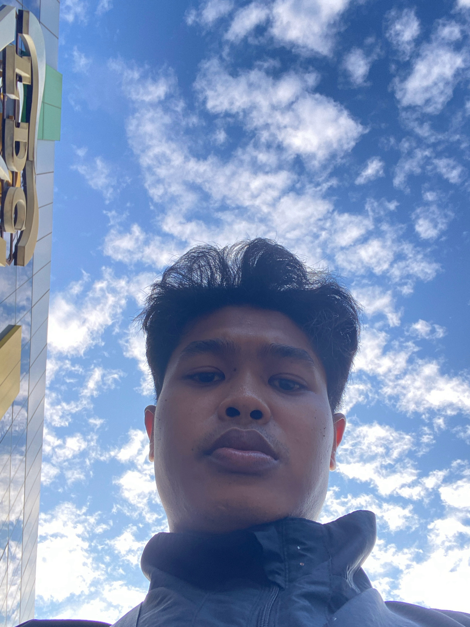 The image is a low-angle, selfie-style shot taken outdoors. In the foreground, a young person's face is prominently featured, looking directly at the camera. They have dark, somewhat messy hair and are wearing a dark jacket. The background is dominated by a bright blue sky filled with scattered white clouds. To the left of the frame, a portion of a modern building with a glass facade and a large, metallic architectural element is visible. The architectural element appears to be a stylized letter or symbol. The overall impression is that the photo was taken in an urban environment, possibly in Batam, Indonesia, given the building's modern aesthetic and the clear sky.