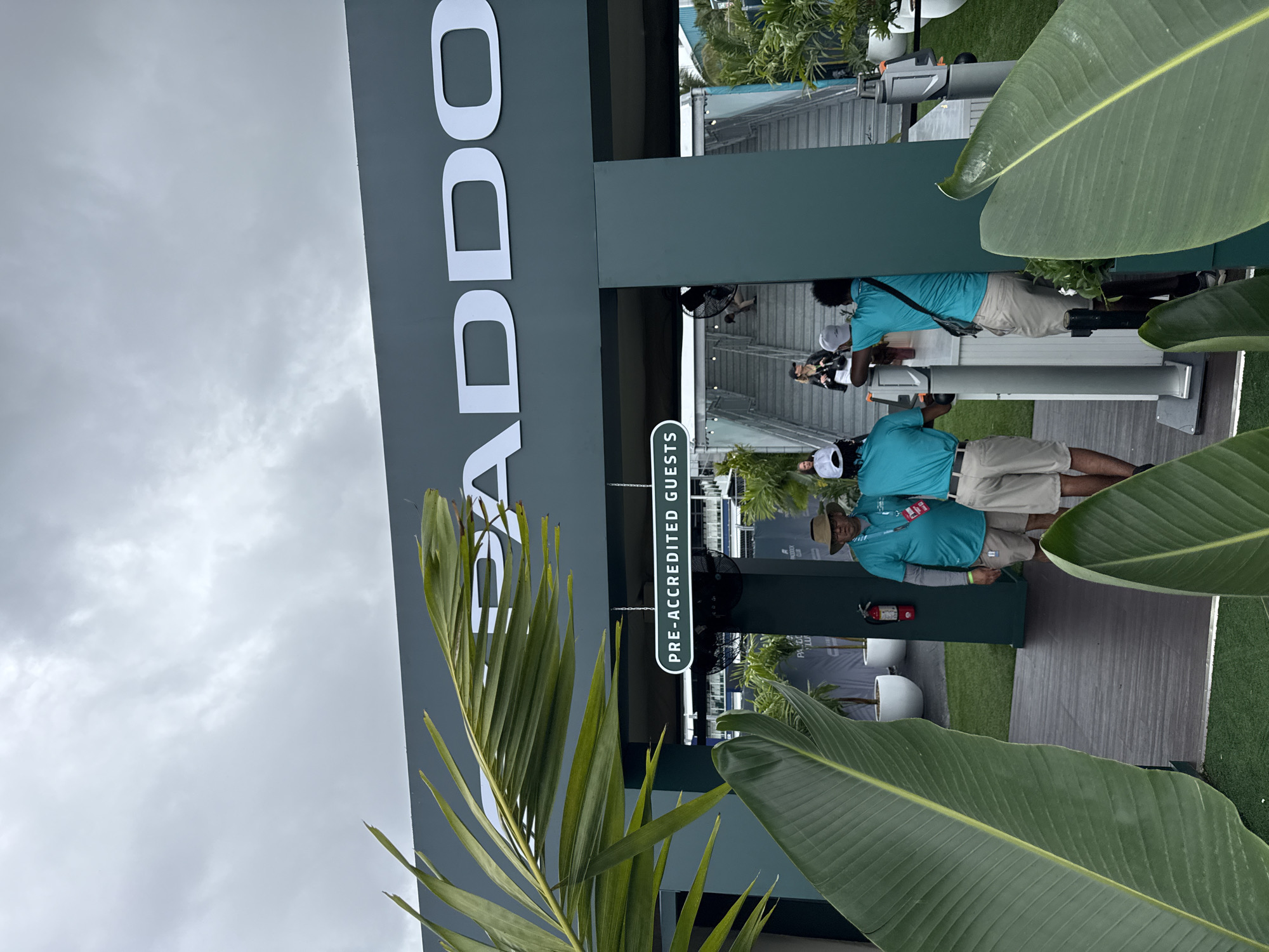 The image shows an outdoor scene at what appears to be an event entrance in Miami Gardens, Florida. A large sign with the word "PADDOCK" in white letters is prominently displayed on a dark gray or green background. A sign below reads "PRE-ACCREDITED GUESTS". Two individuals wearing light-colored shorts and blue shirts are positioned behind a desk-like structure; they may be checking credentials. One of them wears a wide-brimmed hat. Various potted plants, including palms and banana-like trees, are scattered around the area, contributing to a tropical atmosphere. The sky is overcast. The location is suggested by the presence of a Paddock Club, common at auto racing circuits such as the Miami International Autodrome.