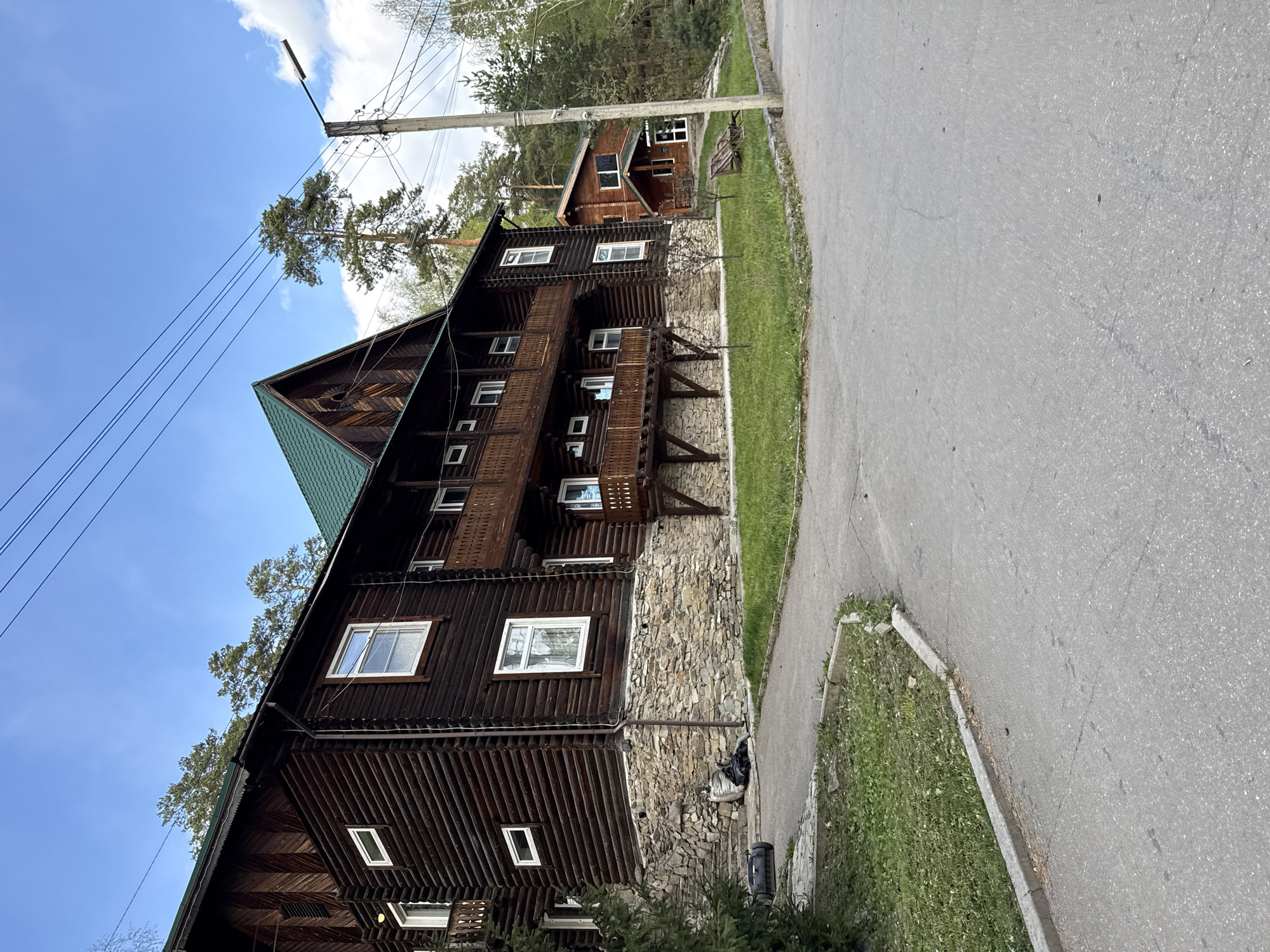 The image depicts a multi-story wooden building with a stone foundation, likely a hotel or lodge, located in Zolotoi Plyazh, Russia. The building features a green roof, multiple windows, and balconies with wooden railings. A utility pole with wires is visible in the foreground, and a paved road runs alongside the building. A smaller wooden structure is seen further in the background, nestled amongst trees. The sky is blue with scattered clouds, indicating a sunny day.