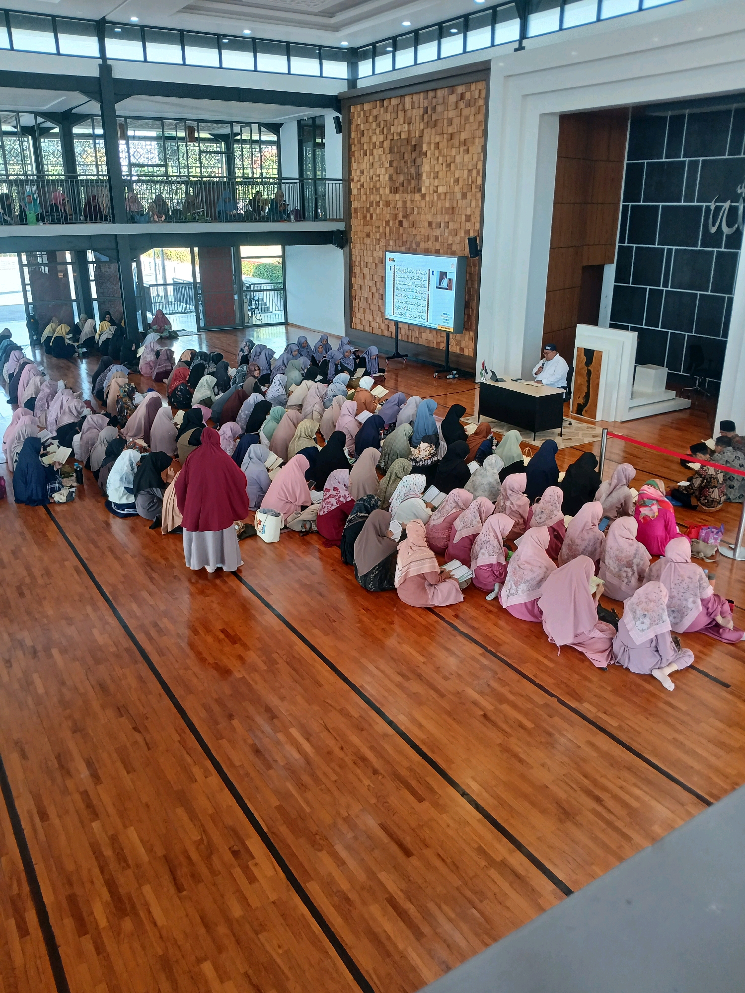 A high-angle, full shot captures an indoor scene featuring a large gathering of people, predominantly women wearing hijabs, seated on a polished wooden floor. The individuals are positioned in rows, facing a presentation screen and a man speaking at a lectern. The setting appears to be a communal hall or institution, possibly a mosque or educational facility, with multi-level architecture visible in the background, featuring glass railings and decorative wooden paneling.
The presentation screen displays text and an image, suggesting an educational or religious discourse. The speaker, a man in a white shirt and cap, is seated at a dark lectern, holding a microphone. The overall atmosphere seems formal and focused, with individuals engaged in listening or reading from books or pamphlets in front of them. The flooring is a warm, varnished wood with black inlaid lines, resembling a basketball court. Natural light enters from large windows and openings, illuminating the space. The location is identified as Arjasari, Indonesia.