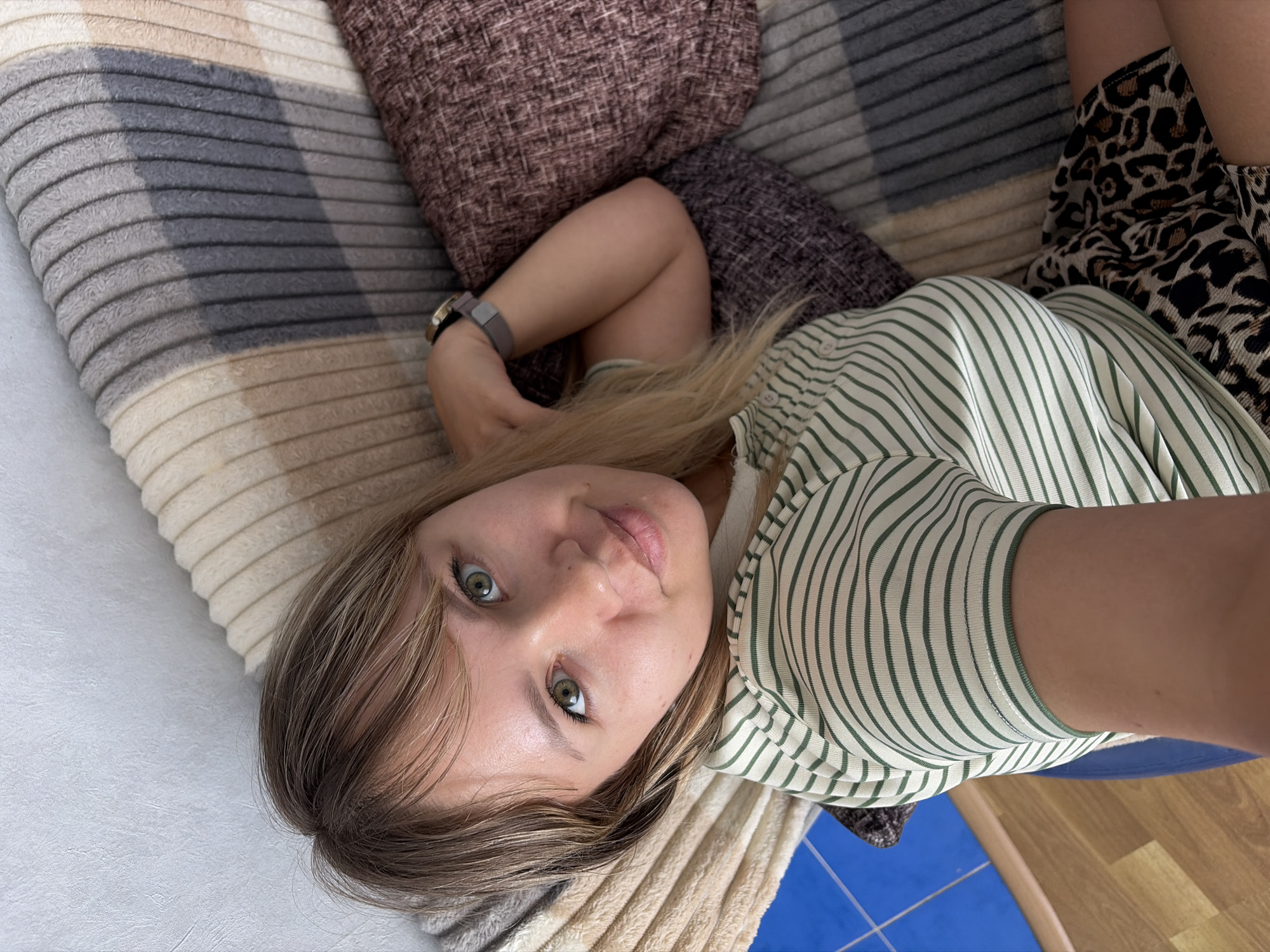 A young woman with light brown hair and prominent green eyes gazes directly at the camera in a close-up, slightly overhead shot, suggestive of a selfie. She is lying down, her head resting on a textured brown pillow, and her body on a cozy blanket with broad horizontal stripes in shades of beige, gray, and light brown. She wears a light-colored shirt adorned with thin dark green horizontal stripes and a skirt or shorts featuring a bold leopard print pattern. A watch or fitness tracker with a gray band and a metallic case is visible on her left wrist, which is positioned above her head. Her expression is calm and open, with a hint of a gentle smile. The lighting appears bright and natural, indicating an indoor, daytime setting. In the lower right corner, a segment of a blue mat and a light-colored wooden floor are visible. There is no visible text in the image. The scene depicts a casual moment of relaxation in Chaikovsky, Russia.