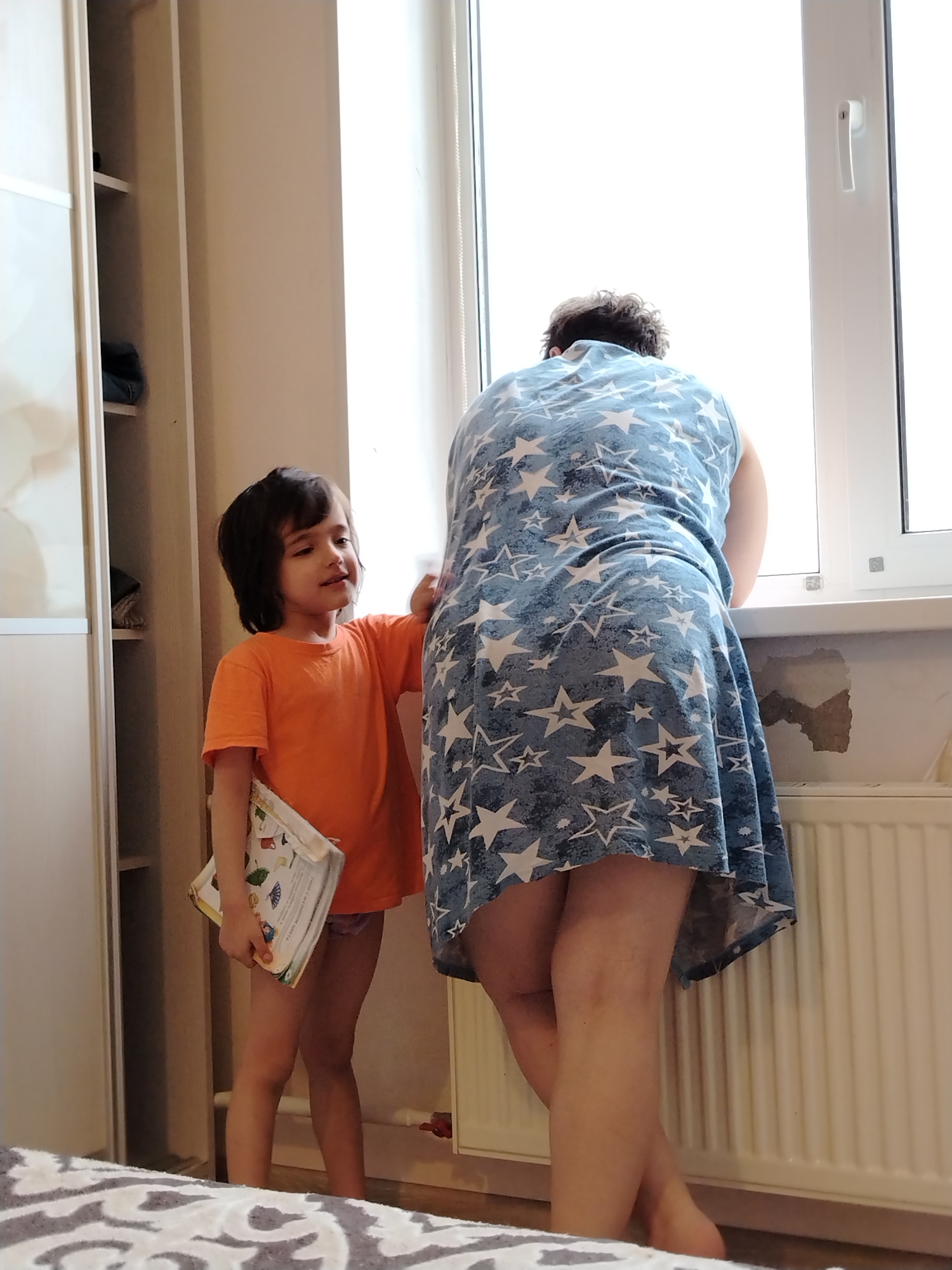 A young child stands to the left of an adult female in a room, possibly in Noviy, Russia, given the prompt. The child, likely a boy, wears an orange t-shirt and carries a book under their left arm. The adult female, with dark curly hair, is facing away from the camera, looking out a window. She is wearing a blue dress with white star patterns. The room appears to be a bedroom, with a light-colored wardrobe to the left and a radiator under the window. The wall below the window shows signs of discoloration or damage. A bed with a patterned bedspread is partially visible in the foreground.