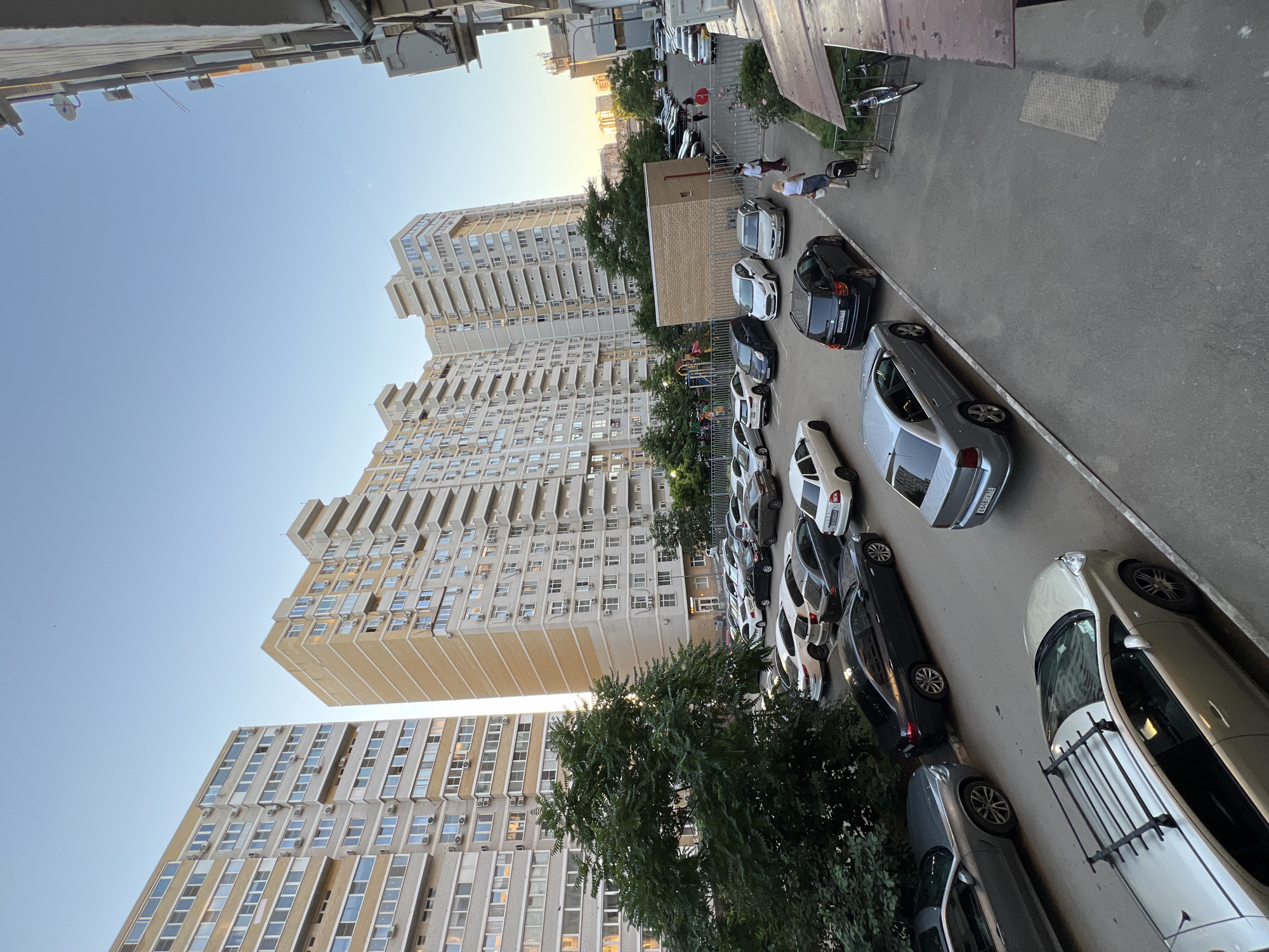 The image depicts a residential courtyard in the city of Krasnodar, Russia, likely during late afternoon or early evening, indicated by the soft, warm light and long shadows. The scene is dominated by several tall, modern apartment buildings, characteristic of urban development in Russia, with beige and light-gray facades, numerous balconies, and external air conditioning units visible on many windows.
In the foreground and mid-ground, a densely packed parking lot is filled with a variety of passenger cars, predominantly sedans and SUVs, in various colors including silver, black, white, and dark gray. Some cars have Russian license plates visible. The pavement is asphalt.
Several people are present. Two women are walking away from the viewer in the mid-ground, one in a white top and blue skirt, the other in a burgundy top and dark trousers, each carrying a bag. Further back, near a playground area with colorful structures, a few other figures are indistinctly visible, possibly children or adults.
A large, mature green tree with dense foliage is situated on the left side of the frame, partially obscuring one of the apartment buildings and casting shadows onto the parking area. Adjacent to the parking lot, a metal fence separates it from a small, brick-clad building and a children's play area. On the far right, a section of another building facade is visible, with windows, an air conditioning unit, and a security camera. A bicycle is parked in a rack near the edge of the frame on the right.
The sky is clear and blue, with a subtle gradient towards a lighter hue near the horizon, suggesting the sun is low. The overall impression is of a typical urban residential setting in Russia, focused on functionality and community living.