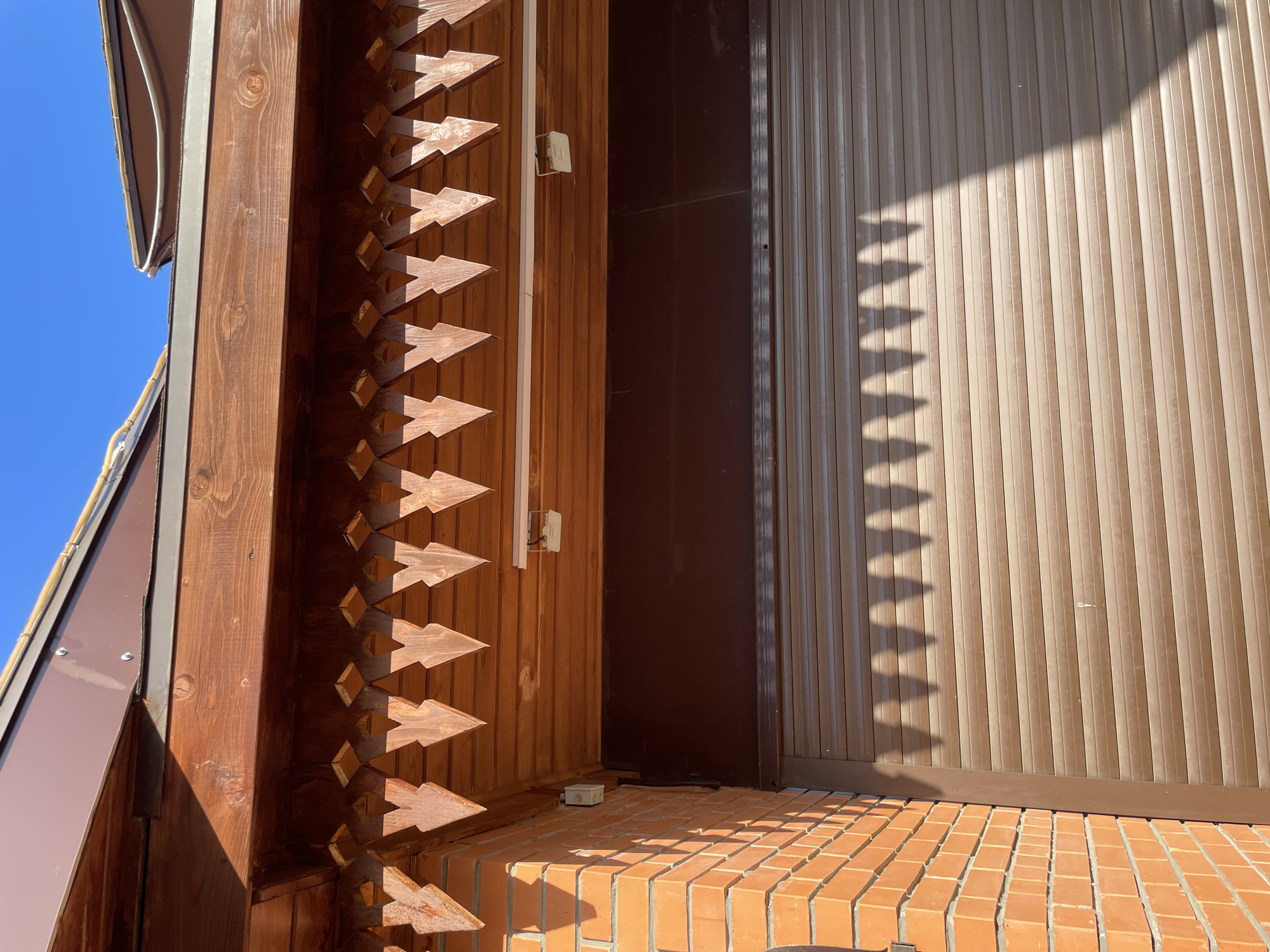 The image shows a corner of a building, likely a house, with a combination of wood, metal siding, and brick construction. The wooden section is decorated with a distinctive, arrow-shaped pattern along the edge. Metal siding in a brown hue is visible, creating a vertical lined pattern. A brick platform or stoop is in the foreground. A shadow mimicking the arrow-shaped design on the wood falls on the siding. Part of a blue sky is visible in the background. There is no evidence to determine the location to be the city of Izhevsk, Russia.