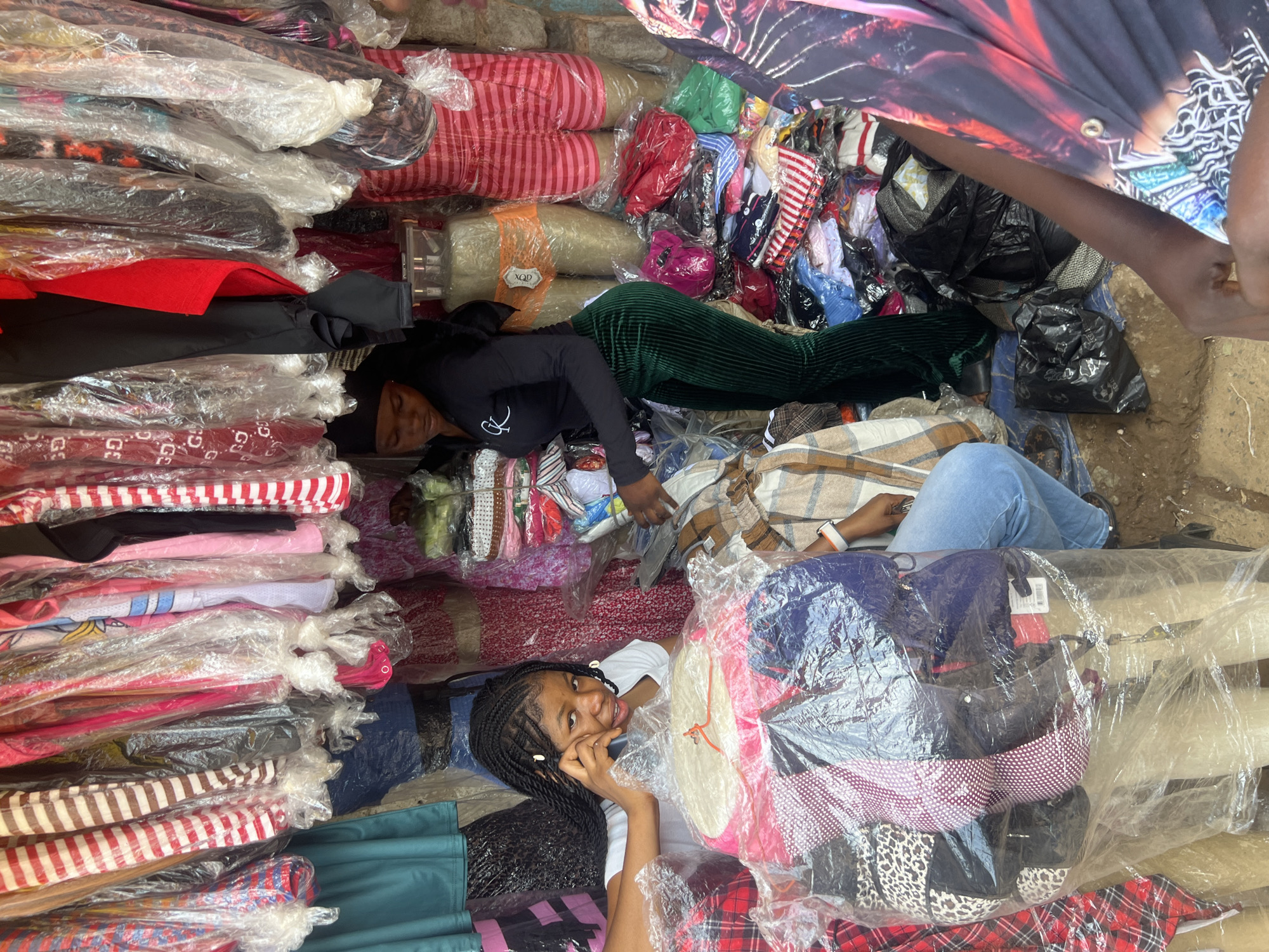 A high-angle shot depicts an outdoor market scene in Unknown, Unknown, filled with packed merchandise. At the center bottom, a young Black woman with braided hair smiles and holds a cell phone to her ear. Another Black woman is located near the top center of the image, wearing a long-sleeved black shirt and green pants. She appears to be surrounded by merchandise as well. The market is full of stacks of clothing items, many wrapped in plastic, in a variety of colors and patterns, and black plastic bags. A person wearing blue jeans is also located at the center-right of the image. A figure with darker skin is located at the top right corner. The ground is visible in some areas, revealing dirt or concrete.