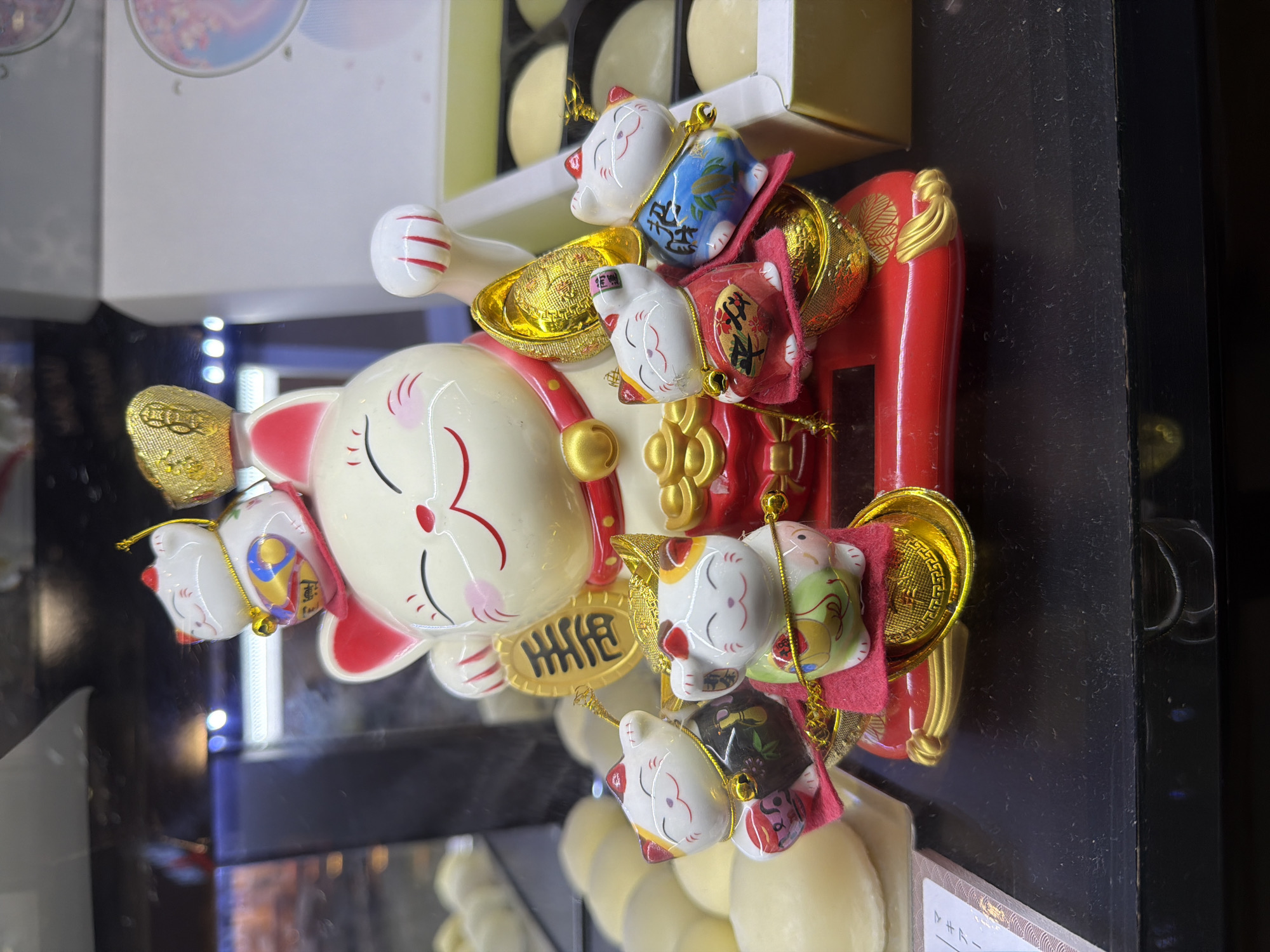 A Maneki-neko, or lucky cat figurine, sits on a black surface, likely a display shelf. The main cat is large and white with pink accents on its ears, nose, and paws, and is adorned with a red collar and gold accessories, including a gold coin and a gold ingot. It holds a gold coin in its left paw. Several smaller Maneki-neko figurines surround the base of the larger one, each with its own unique colorful attire and accessories, including gold coins and bells. The figurines are displayed in what appears to be a retail setting, possibly a gift shop or a store selling Asian cultural items. In the background, blurred lights and shelves suggest the interior of a commercial establishment. The presence of a few white, round objects in the background could be pastries or other merchandise. The image does not provide any specific visual cues to pinpoint its location in Da Nang, Vietnam.