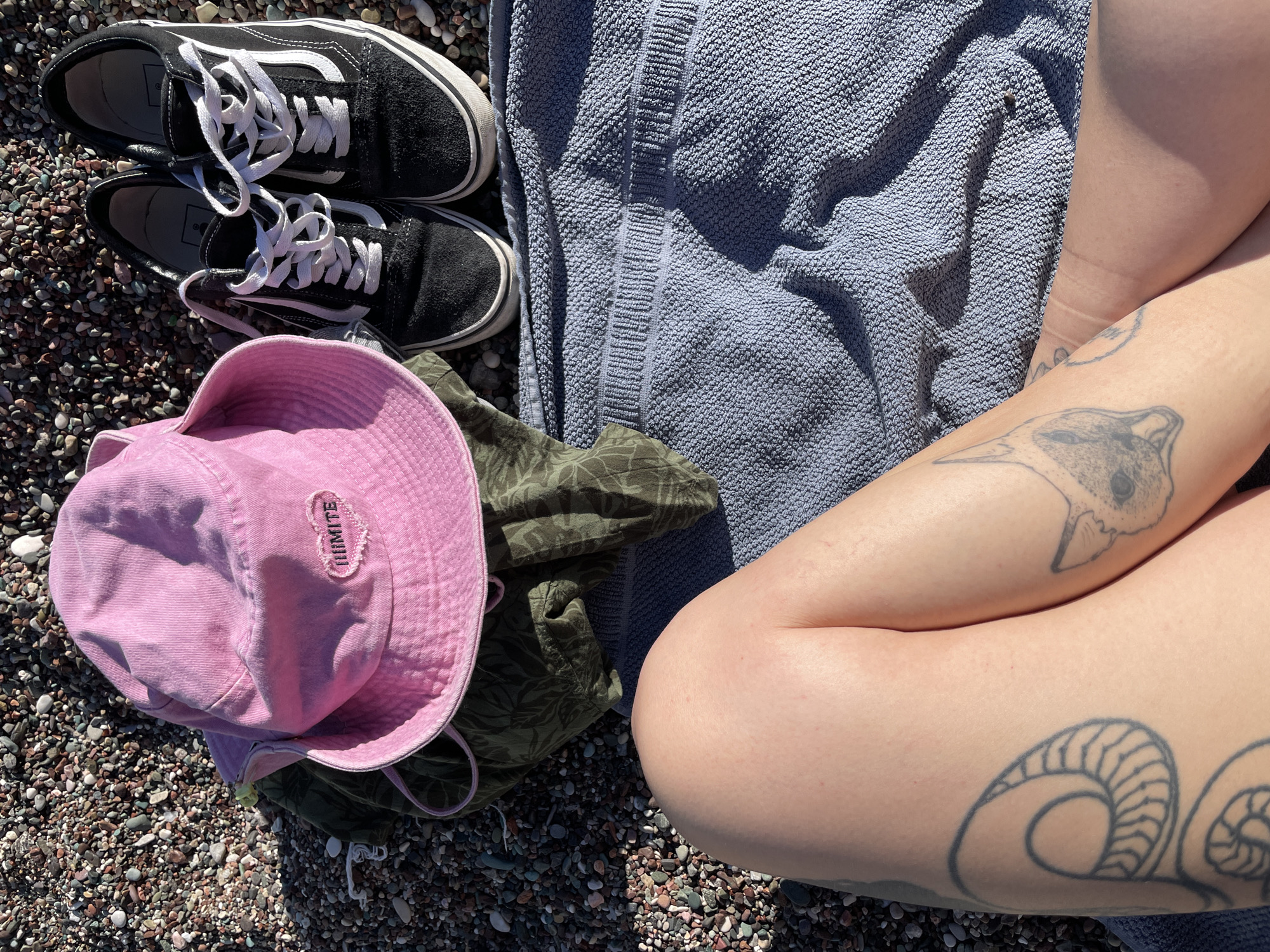 A ground-level shot shows a person seated on a pebble beach near Rafailovići, Montenegro. Their legs are crossed on a blue textured towel. The person has tattoos on their legs, one depicting a fox and another, a stylized ram’s horn. Adjacent to the towel are a pair of black Vans sneakers. At the left of the frame, on the pebbles, is a pink bucket hat and a green patterned shirt. The ground is made of small pebbles of various colors. Sunlight illuminates the scene.