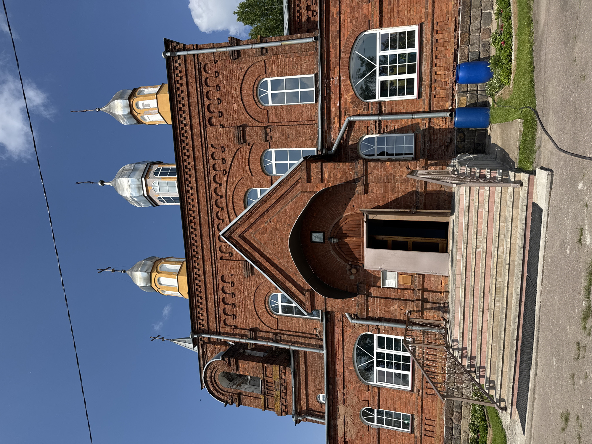 This image depicts the front facade of a red brick church, likely located in Chudovo, Russia, under a clear blue sky with scattered clouds. The church features a bell tower on the left, a central entrance with a large arched doorway and wooden doors, and three onion-shaped domes topped with crosses on the roof. The architecture is characteristic of Eastern Orthodox churches, with ornate brickwork and arched windows.
The entrance is accessed by a wide set of stairs with red risers and stone treads, flanked by metal railings. There is a dark mat at the bottom of the stairs. To the right of the entrance, two large blue plastic barrels are visible, situated on a paved area near a stone wall. Green grass and some flowering plants are growing in the foreground and to the sides of the church. A black hose lies on the ground in front of the barrels. The overall scene suggests a sunny day in a rural or semi-urban setting. There are no people visible in the image. The perspective is from ground level, looking slightly upwards at the church.