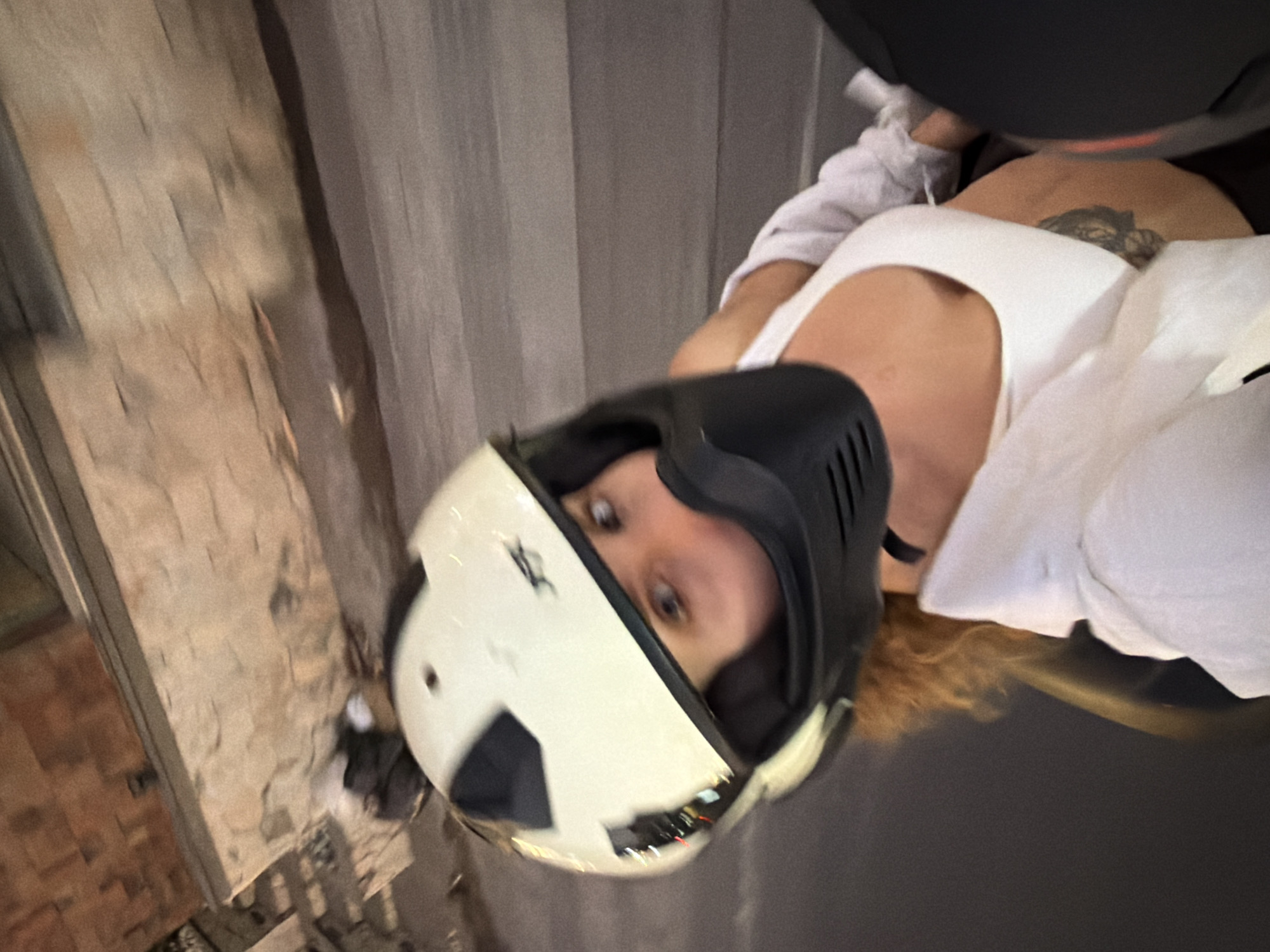 A person, likely female, is depicted from a high-angle perspective, positioned as a passenger on a moving vehicle, presumed to be a motorcycle or scooter, at night in North Kuta, Indonesia. She is wearing a light-colored full-face motorcycle helmet with a black chin bar, which partially obscures her face, revealing only her eyes, which are directed towards the camera. Light brown, curly hair extends from beneath the helmet. She is dressed in a white tank top, overlaid with a loose white long-sleeved shirt. A dark, intricate tattoo is visible on her right side, below the rib cage. The background exhibits substantial motion blur, indicative of movement. An outdoor brick or stone wall with a defined ledge is visible in the upper left, while a blurred road surface occupies the lower right. Reflections of indistinct urban lights are visible on the helmet's surface.