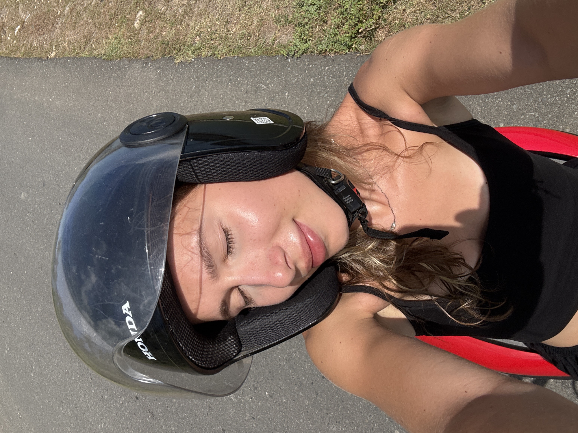 This is a close-up, overhead, portrait-orientation selfie of a young woman wearing a black helmet with a clear visor. She is leaning her head back, with her eyes closed and a gentle smile on her lips. Her hair, a mix of blonde and brown, is partially visible around the helmet. She is wearing a black tank top. A small silver necklace is visible around her neck. The helmet has the "HONDA" logo on the side. The scene appears to be outdoors, with a paved surface, possibly a road or path, in the background. There is dry grass and some greenery visible beyond the paved area, suggesting a warm, sunny environment. The lighting suggests it is daytime, likely mid-morning or afternoon. The overall mood is relaxed and content. The location is Kertasari, Indonesia.