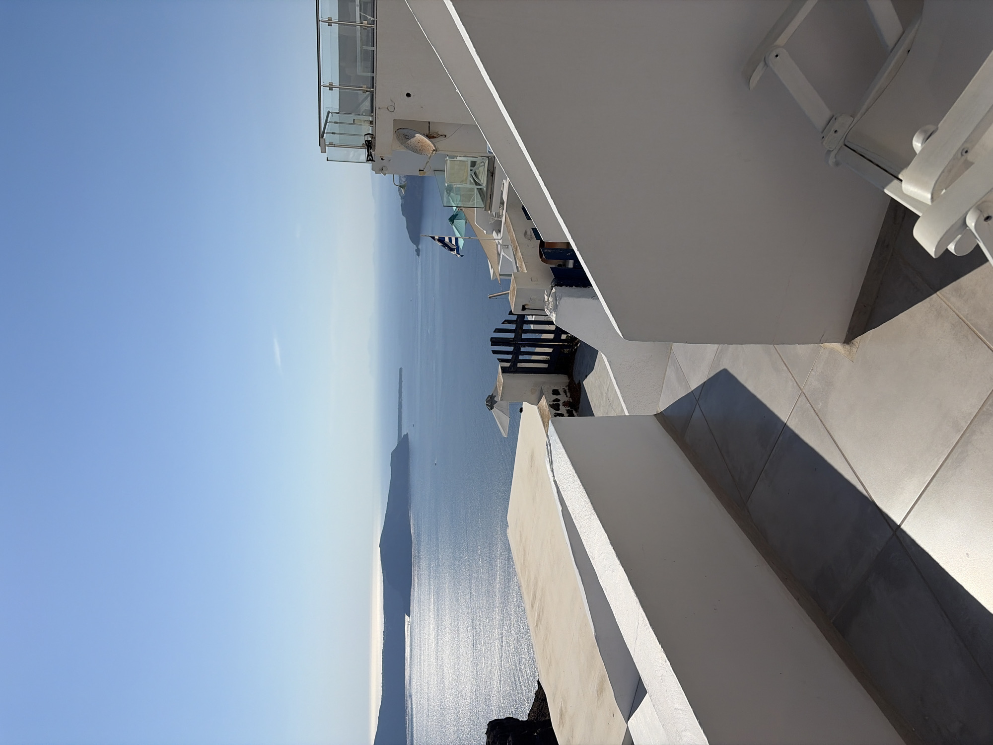 The image depicts a sun-drenched view of the Aegean Sea from an elevated vantage point, likely in Oia or Fira, Thira Municipal Unit, Greece, given the characteristic white-washed architecture and caldera views. In the foreground, a pathway paved with large, square tiles leads towards the sea. To the left, a low, white, stucco wall casts a sharp shadow across the pathway, creating a geometric pattern. A similar white wall rises on the right side, partially obscuring a white folding chair.
Beyond the foreground structures, a multi-level white building dominates the middle ground. A dark blue, slatted gate is visible, with a small blue and white flag flying on a pole above it. A satellite dish is mounted on the exterior wall of this building. Beyond the building, the vast, shimmering expanse of the Aegean Sea stretches towards a distant island chain, bathed in bright sunlight. The sky is a clear, unblemished blue. The scene suggests a tranquil, scenic location, typical of the Cycladic islands.