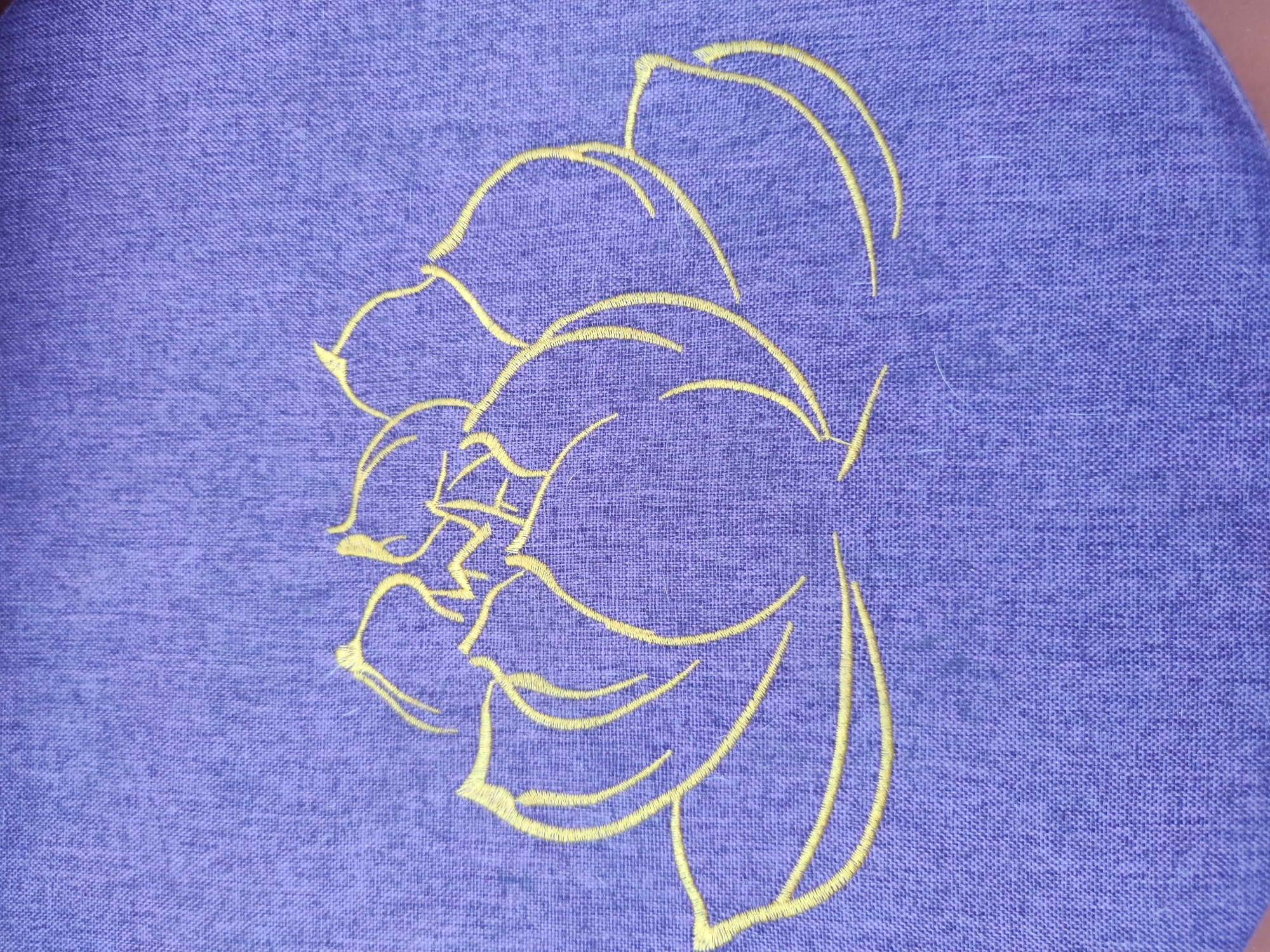 The image shows a close-up of a piece of purple fabric with a yellow embroidered flower design. The design appears to be an outline of a rose, with stylized petals and curves. The fabric has a visible texture.