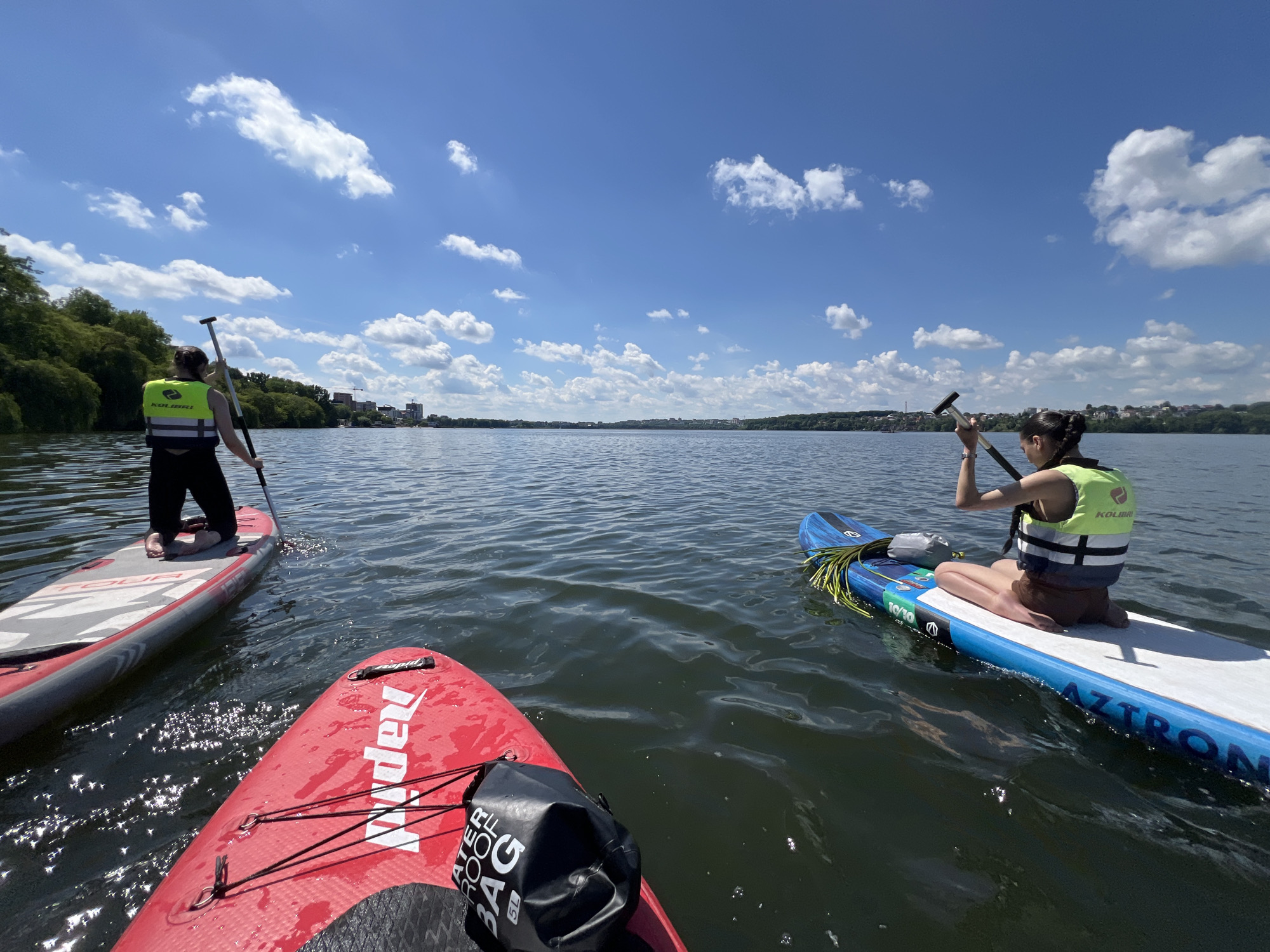 The image shows three people on paddleboards on a body of water, likely Ternopil Lake, in Ternopil, Ukraine. The sky is blue with scattered white clouds. The water is calm with ripples. On the left, a person in a green and black life vest is kneeling on a red paddleboard. In the center, closer to the viewer, is a red paddleboard with a black waterproof bag on it. On the right, another person in a green and white life vest is kneeling on a blue and white paddleboard, holding a paddle. The background shows a shoreline with trees and buildings.