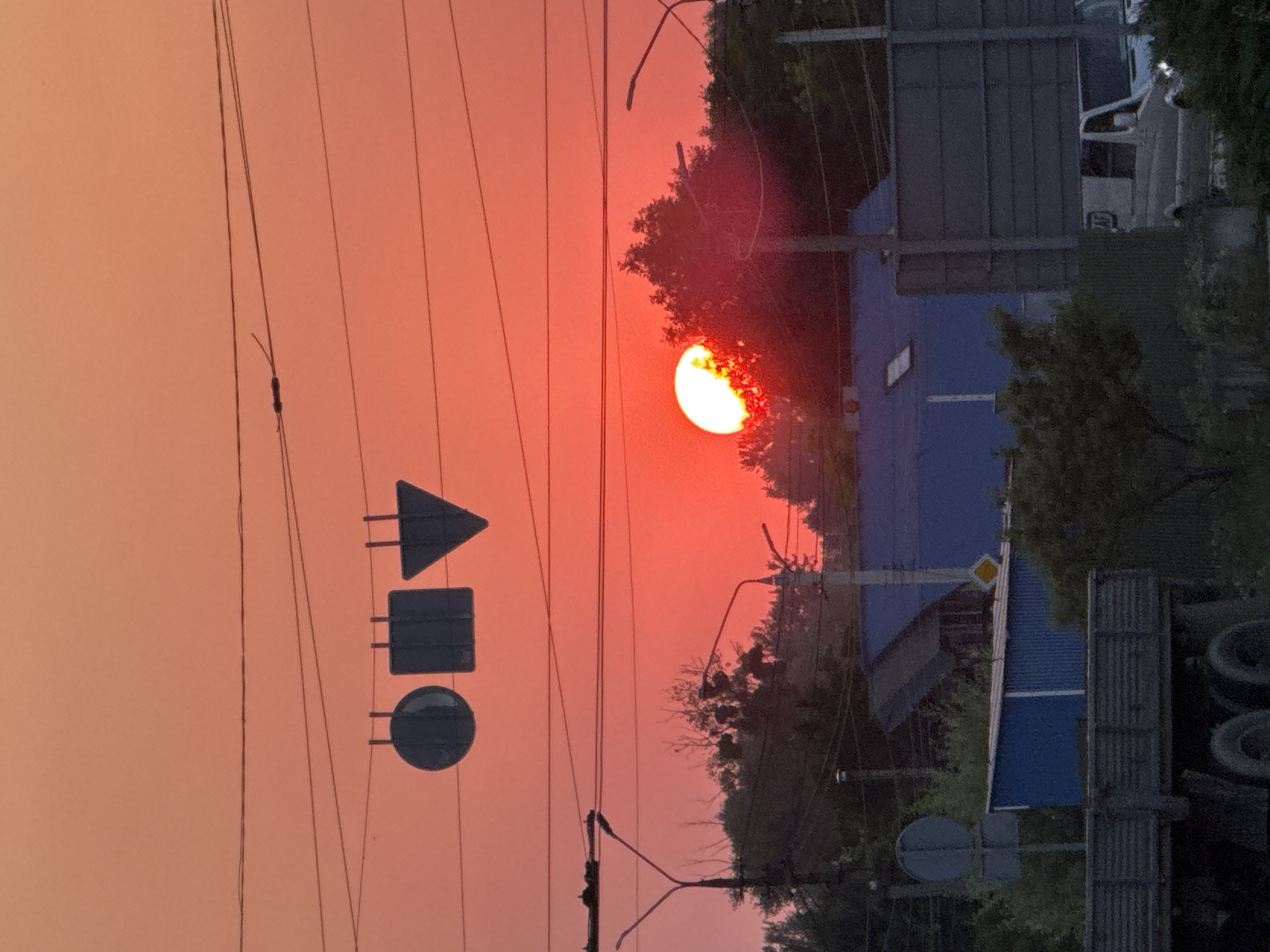 The image captures a vibrant sunset over Chernivtsi, Ukraine, casting a warm, orange-red glow across the sky. Several power lines crisscross the frame, some adorned with traffic signal signs shaped like a circle, square, and triangle. Below these, trees and streetlights create a silhouette against the colorful sky.
In the middle ground, a blue-roofed building with a prominent skylight is visible, partially obscured by greenery. A grey truck with visible tires is parked in the foreground, its flatbed taking up a significant portion of the lower left of the image. To the right, a white van with a visible "AF" logo on its side is parked on a road, its headlights illuminated. The scene appears to be a semi-urban or suburban area, with a mix of residential and industrial elements. No people are visible in the frame.
