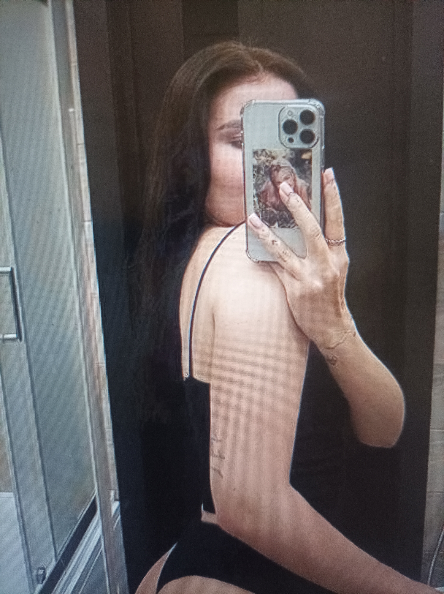 A person is taking a mirror selfie in what appears to be a bathroom or changing room, as indicated by a shower enclosure on the left side of the image. The person is holding a phone with a clear case that displays a picture of a person. The phone case also has a distinctive camera module. The person is wearing a black garment with thin straps, revealing part of their back and arm. They have long, dark hair and are wearing a ring on their finger. There are subtle tattoos visible on their arm. The lighting is artificial, and the overall mood is casual.