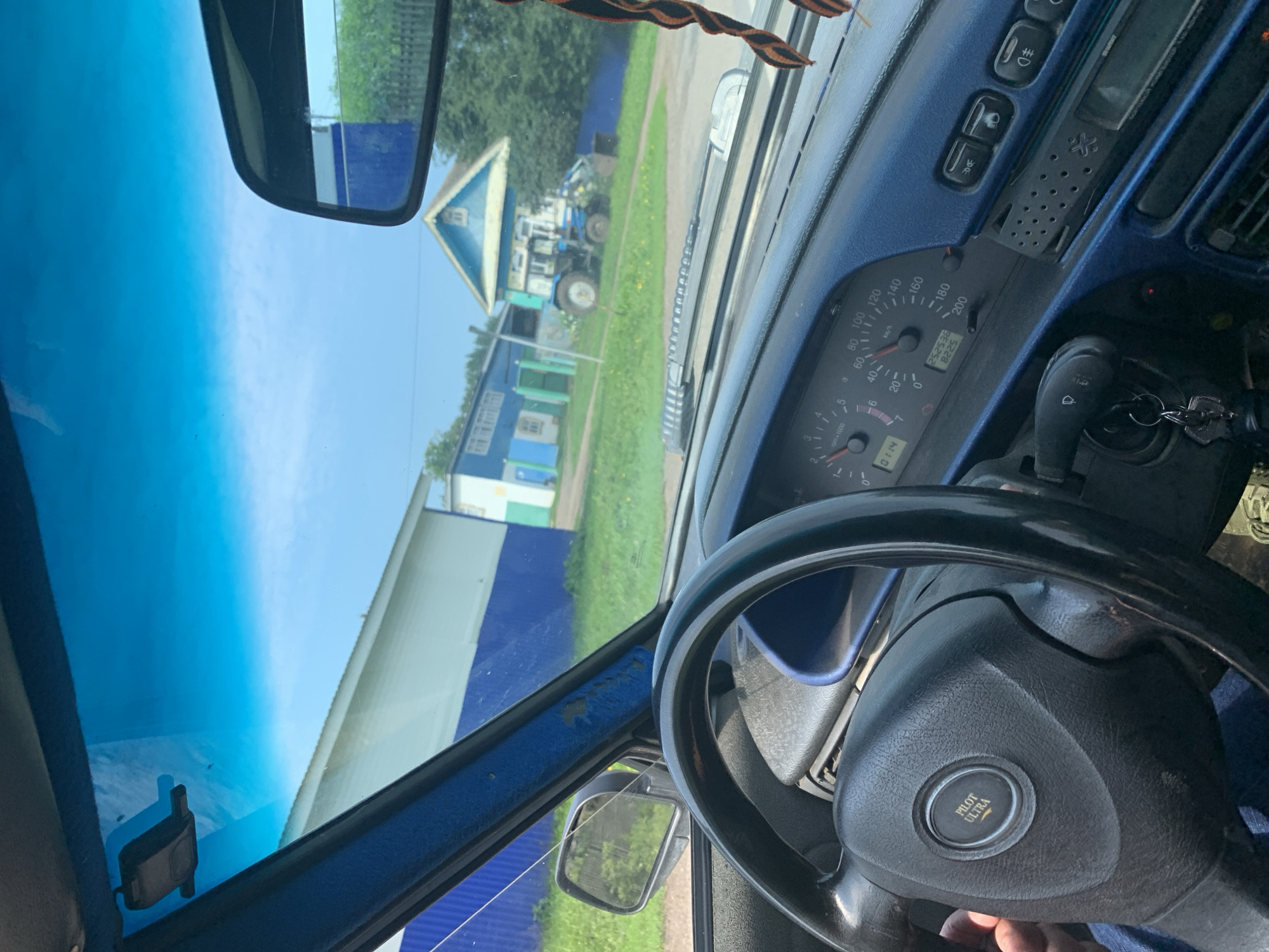 The image is a point-of-view shot from inside a car, looking through the windshield. The car's interior is visible, including the steering wheel with a "PILOT ULTRA" emblem, the dashboard with a speedometer and tachometer, and a blue trim. Outside the car, a rural scene unfolds. Two buildings with blue and white facades are visible, along with a blue tractor. The environment appears to be a village, possibly in Russia, given the architectural style and the presence of a tractor. The sky is clear and blue, suggesting daytime. There are no people clearly visible within the frame, except for a hand holding the steering wheel.