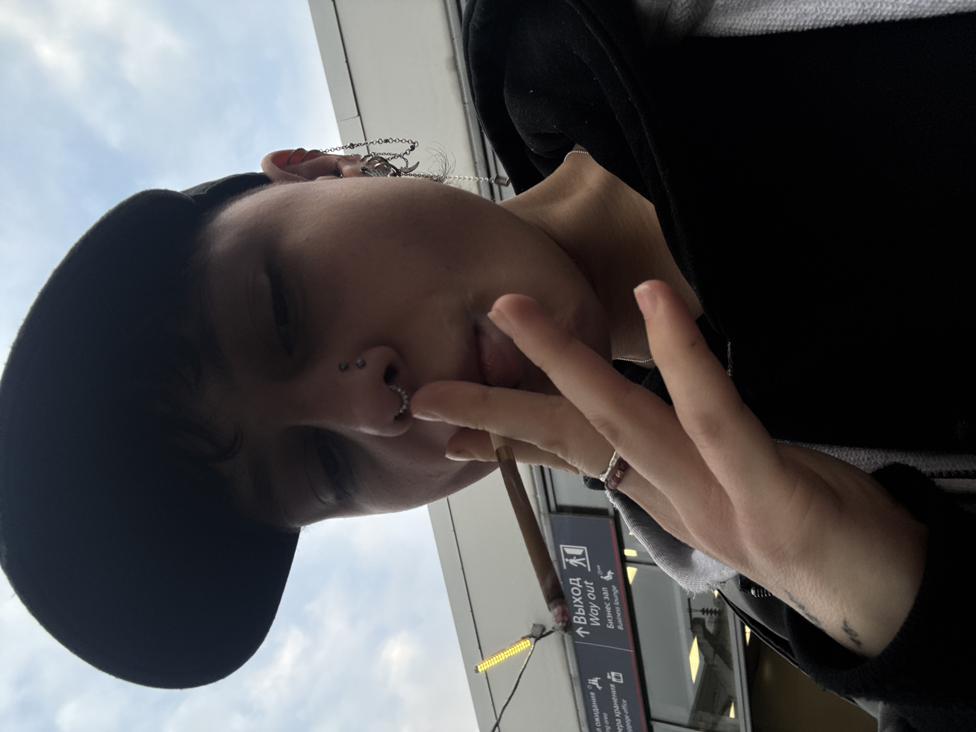 The photograph shows a person wearing a black baseball cap and dark clothing, holding a lit cigarette or joint near their mouth, with their other hand raised in a peace sign. They have piercings in their nose and ears, including decorative chains. The background shows airport signage in both Cyrillic and English that reads, "Выход" and "Way out," with additional text indicating a business lounge and baggage office. This suggests the photo was taken at Pulkovo Airport (LED) in Saint Petersburg, Russia.