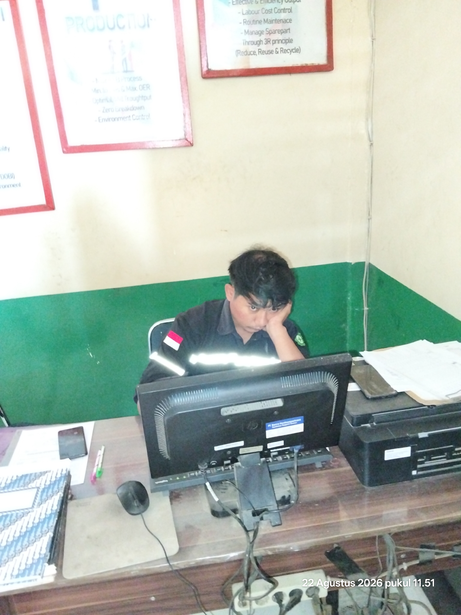 A young man wearing a black uniform with reflective stripes, featuring the Indonesian flag emblem on his left arm, leans his head on his hand while seated at a desk in front of a computer monitor. The room appears to be an office or workstation, with a green and beige painted wall in the background. On the desk, there is a computer monitor, a printer, a mobile phone, pens, a mouse, and a stack of books or documents. A whiteboard or framed poster with text and graphics is visible on the wall to the left. The timestamp "22 Agustus 2026 pukul 11.51" is superimposed on the bottom right of the image. The overall lighting suggests an indoor setting. The city of Unknown, Indonesia, is the stated location.