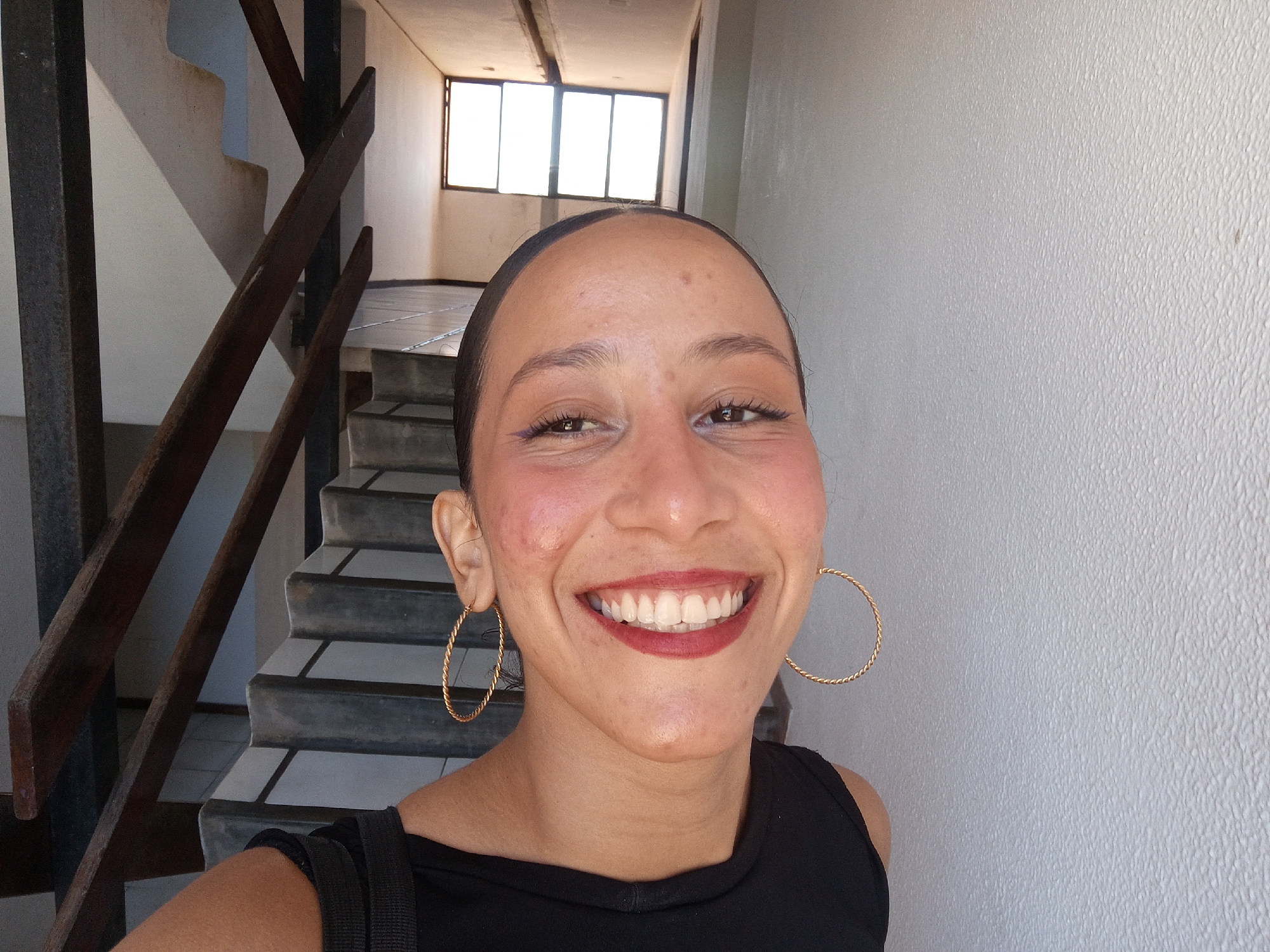 A woman is taking a selfie in a building in Porlamar, Venezuela. She is smiling broadly, showing her teeth, and wearing large gold hoop earrings. Her hair is pulled back, and she has on dark lipstick and eyeliner. She is wearing a black top. Behind her is a staircase with metal railings and light-colored walls. A window can be seen at the top of the stairs, letting in bright sunlight. The wall to her right has a textured surface.